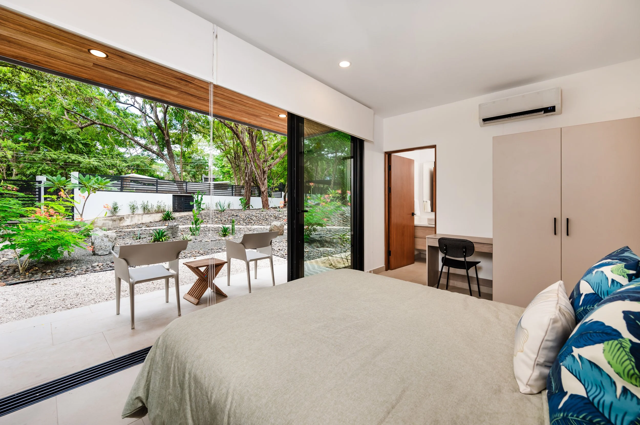 Modern bedroom with sliding glass door opening to a garden patio with trees and plants, observation chairs, and an outdoor landscape.