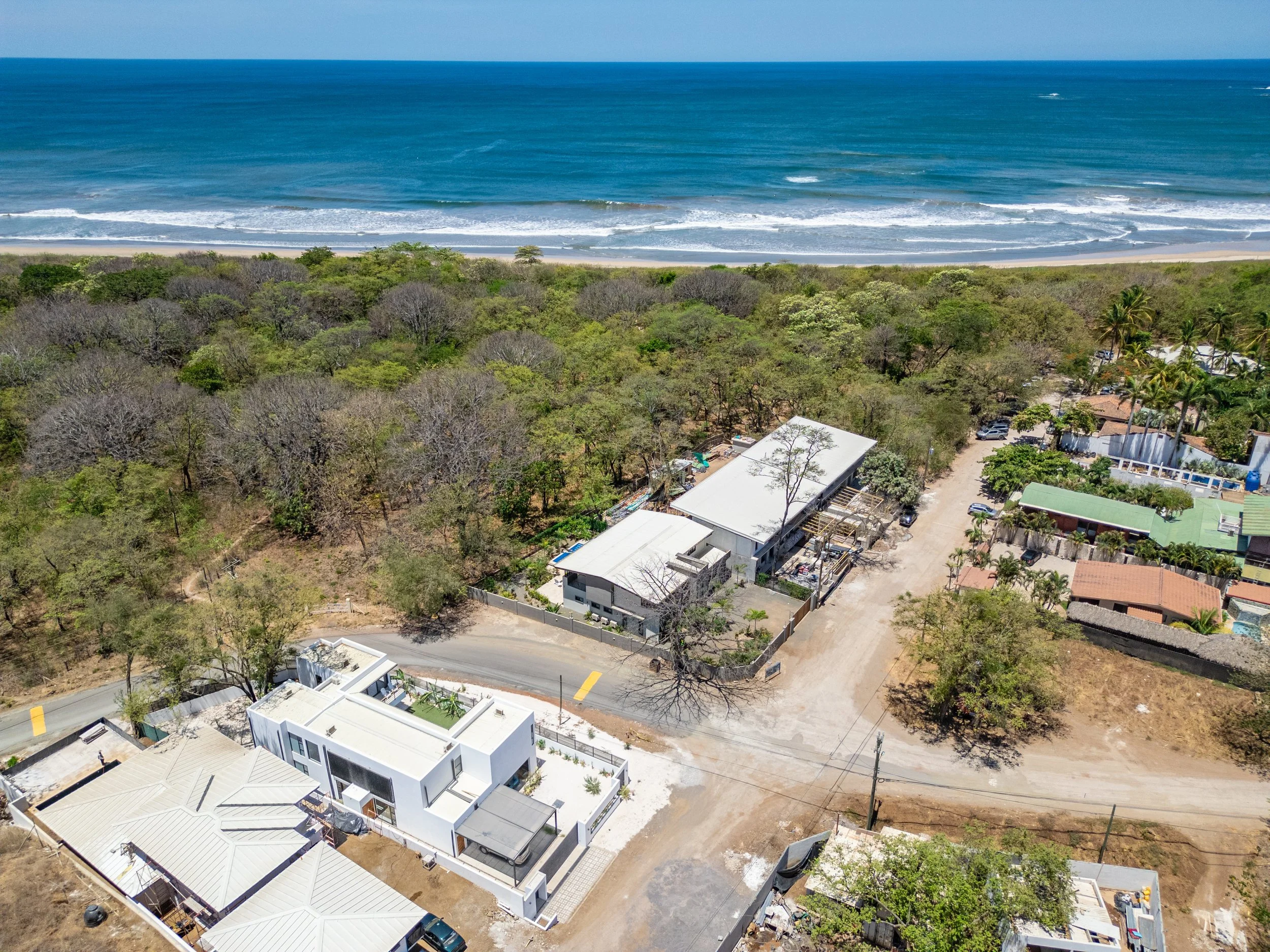 Aerial view of houses near a forest with the ocean in the background, showing clear blue water and white waves.