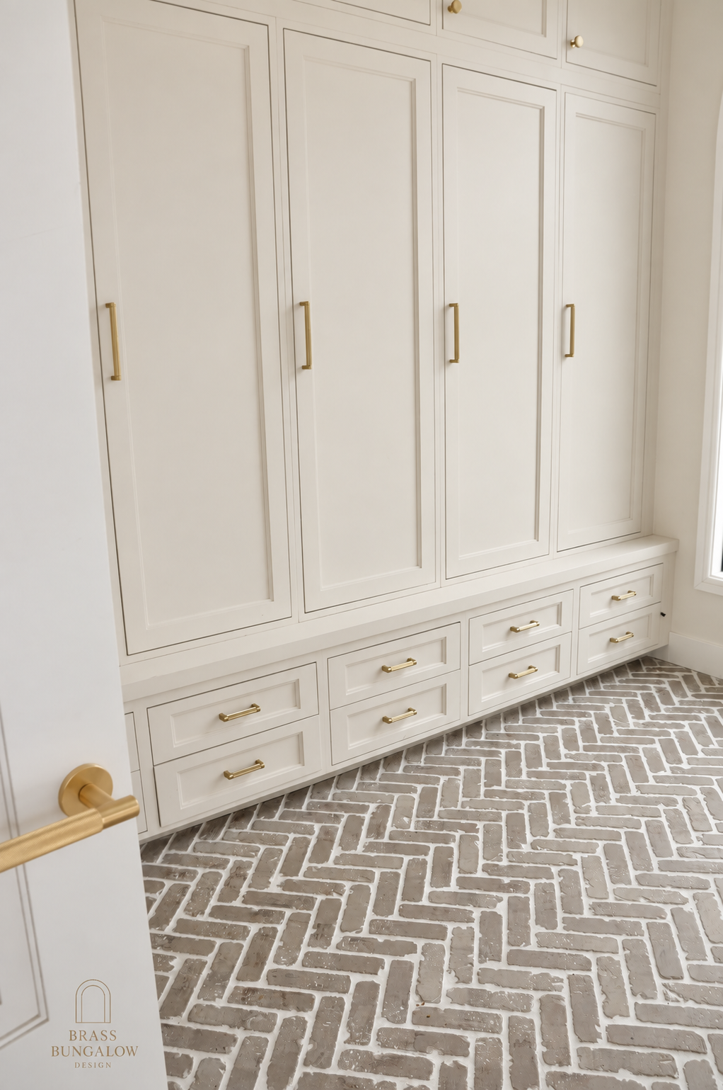 Warm beige mudroom cabinetry with brass hardware and herringbone brick flooring.