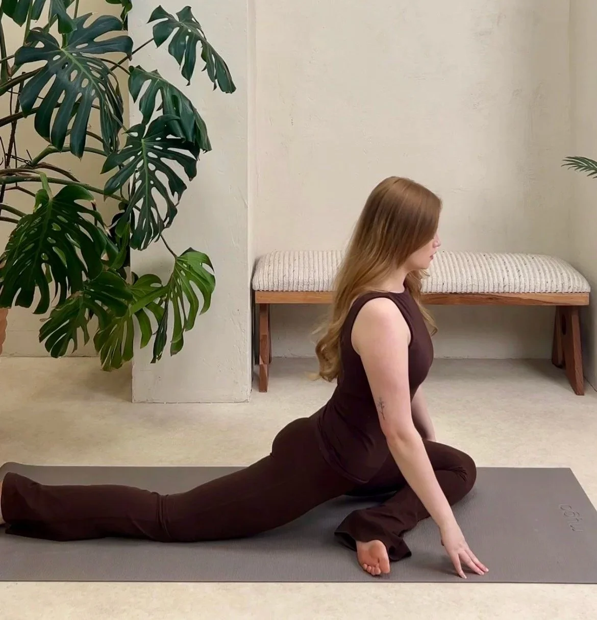 A woman with long red hair practicing yoga indoors, performing a split on a gray yoga mat, with large green plants and a bench in the background.