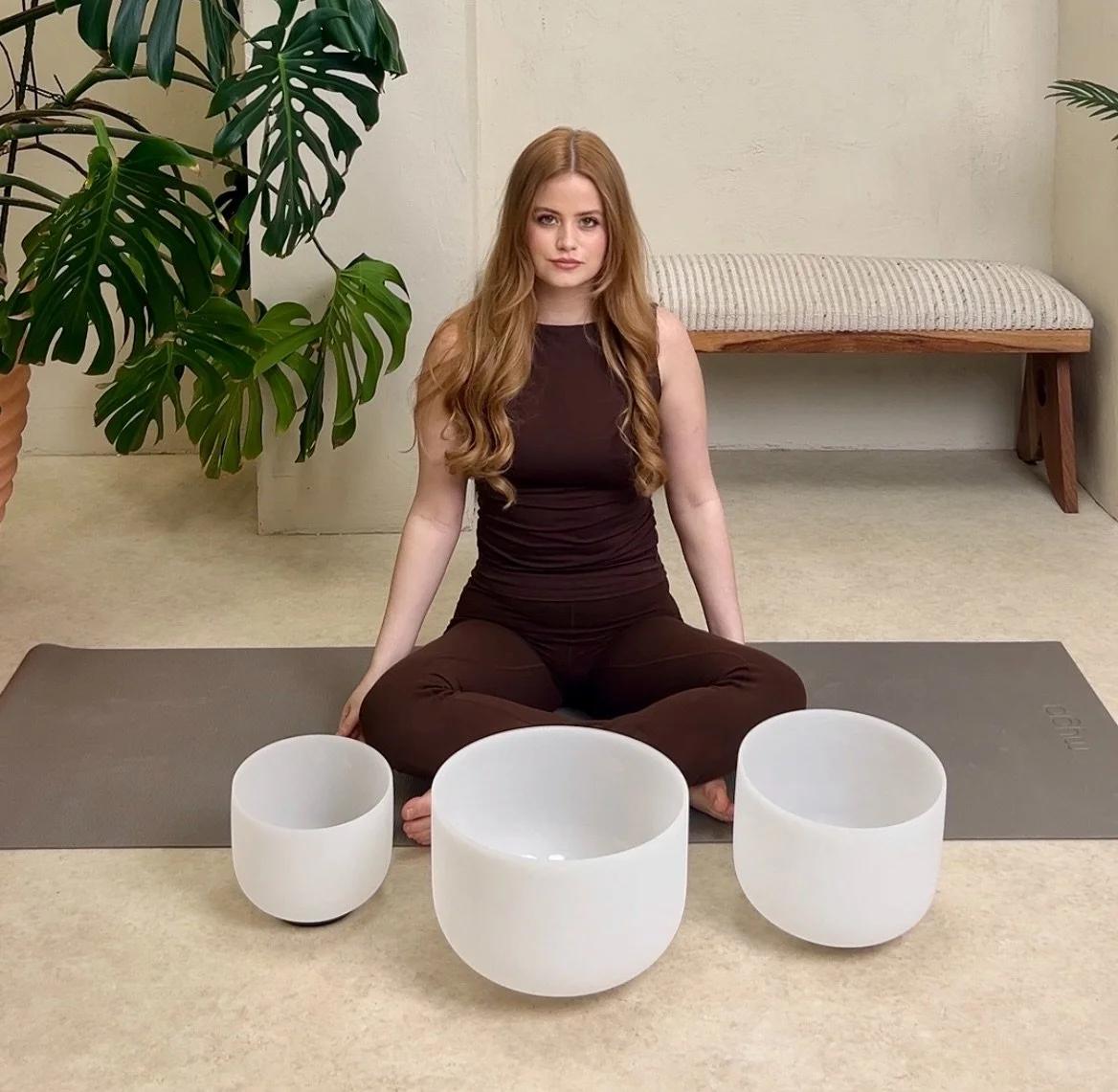 A woman sitting cross-legged on a yoga mat with three white singing bowls in front of her, in a room with a large plant and a bench in the background.