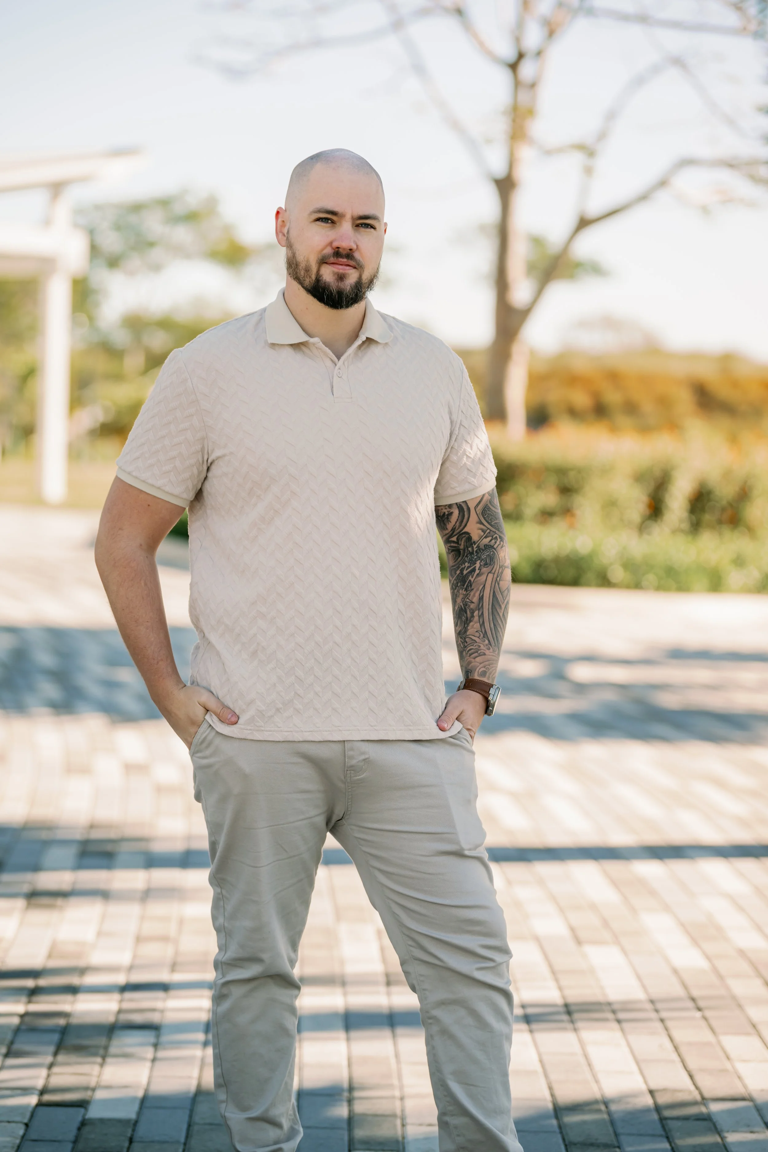 A man with a beard, shaved head, wearing a beige short-sleeve shirt with a diamond pattern and beige pants, standing outdoors on a brick path with trees and greenery in the background during daytime.