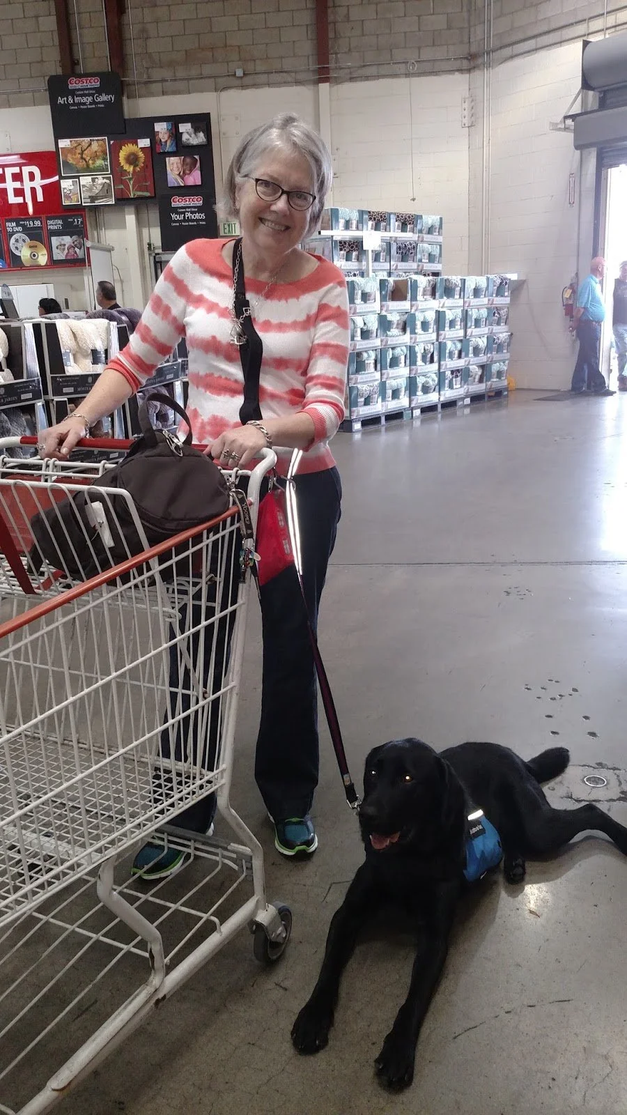 A smiling woman in glasses, a red and white striped shirt, standing with a shopping cart inside a store, with a black dog lying on the floor next to her wearing a service vest.
