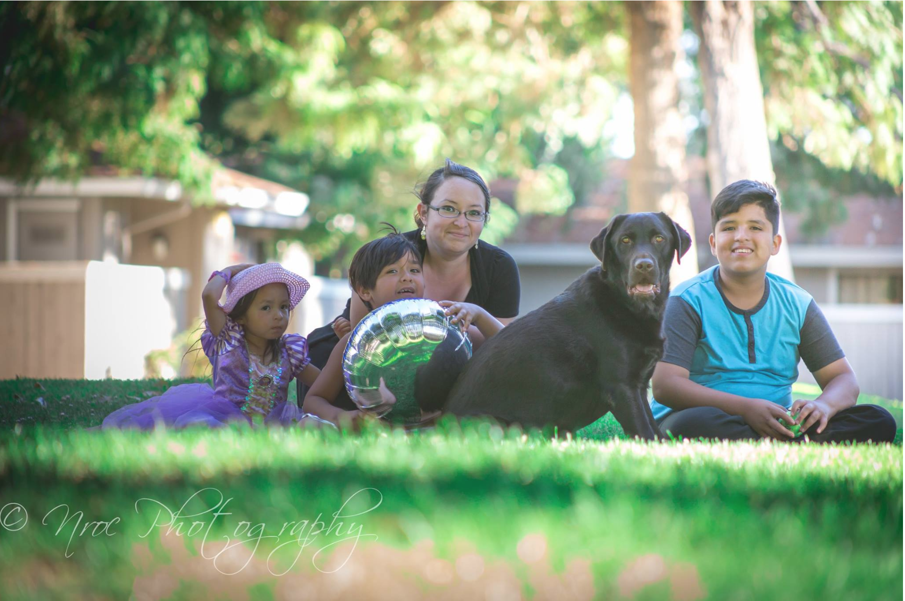 A group of five people and a dog sit on grass in a park with trees in the background. The group includes a woman, two young boys, a young girl in a purple dress and hat, and a black dog. The woman and boys are smiling, the girl and the dog are looking alert, and one boy holds a shiny green balloon.