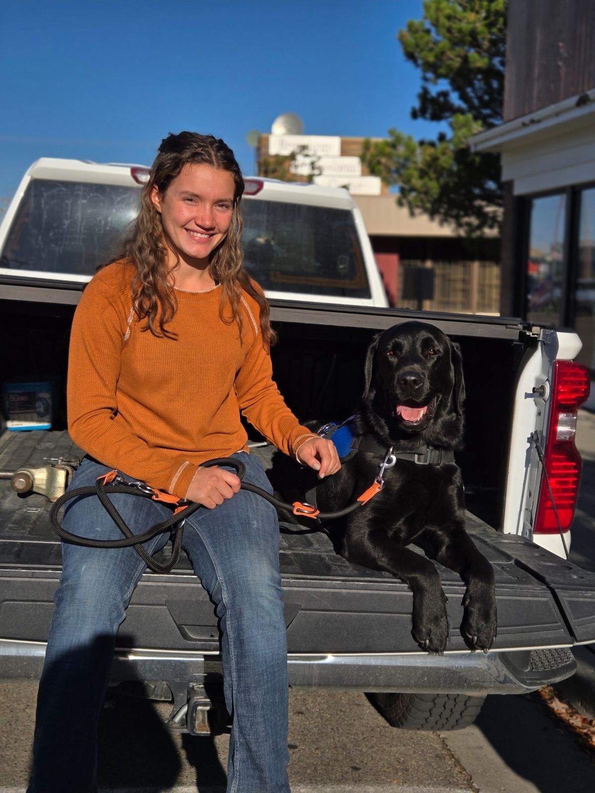 A woman in an orange sweater sitting on the tailgate of a pickup truck, holding a leash attached to a black Labrador retriever dog with a smiling face, in a parking lot on a sunny day.