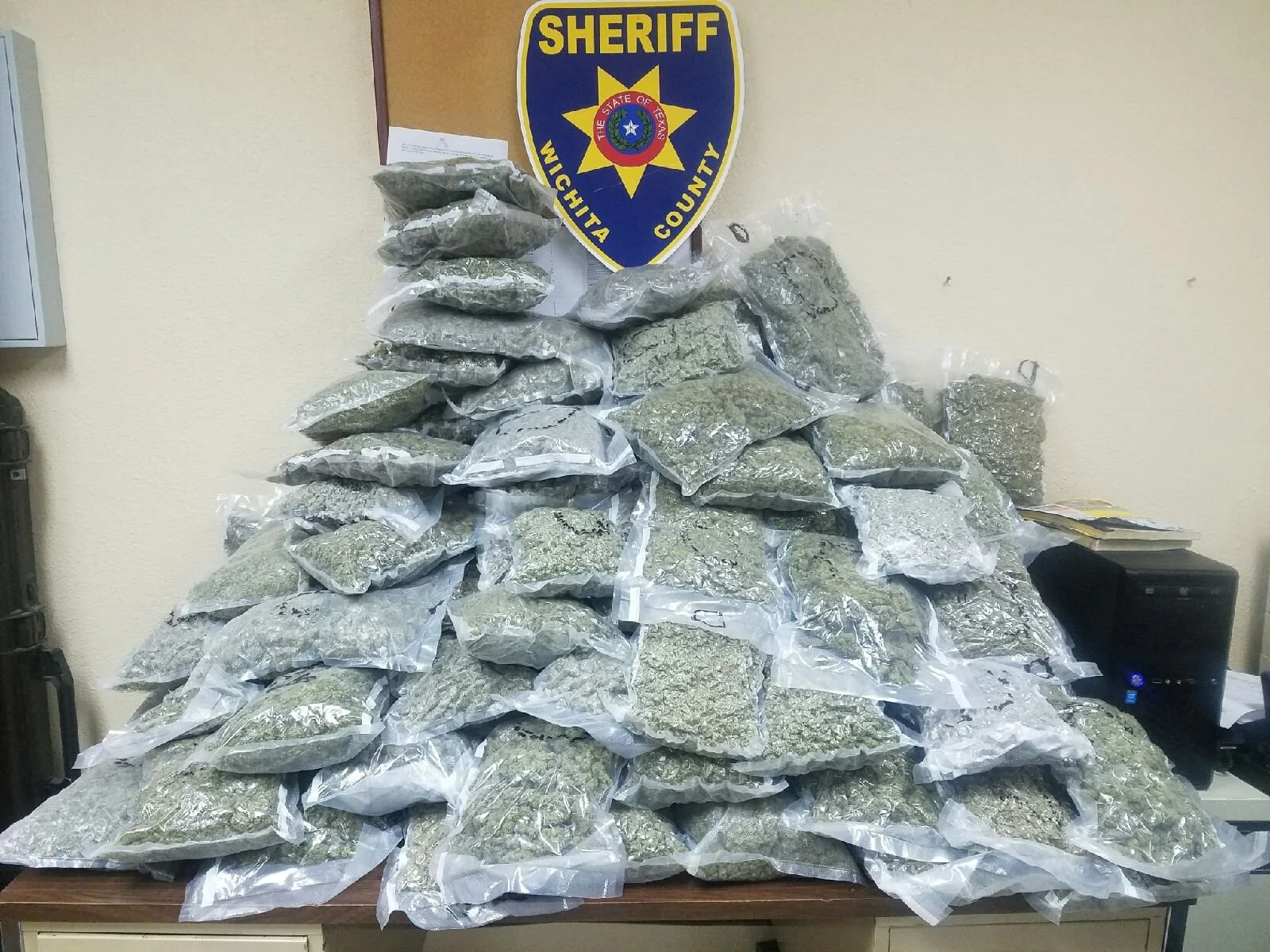 Large pile of plastic bags filled with what appears to be marijuana, with a sheriff's badge on the wall behind it.