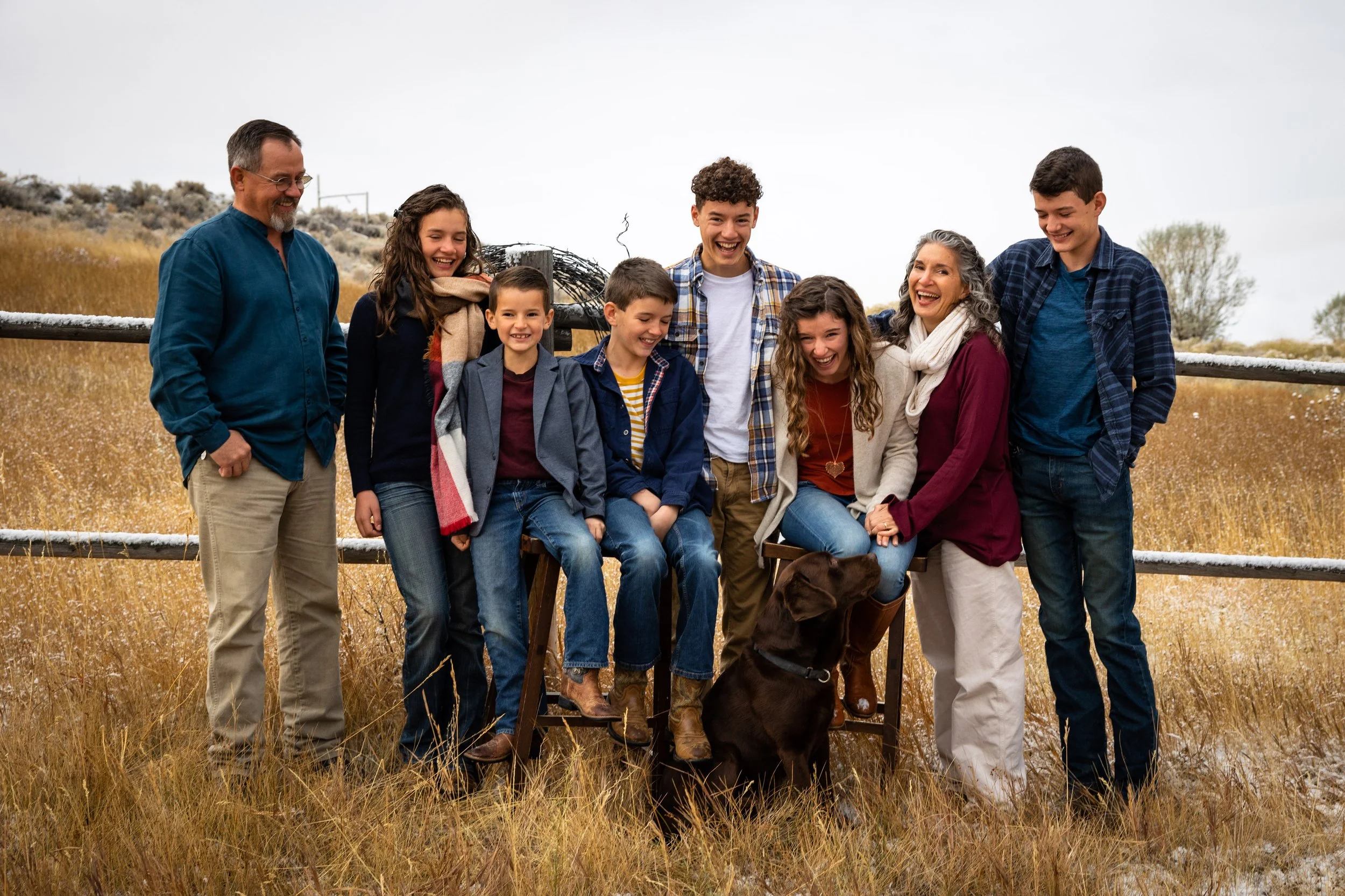 Group of nine people, including children and adults, standing together outdoors on a farm. They are smiling and laughing, with a brown dog sitting in front of them. The background shows a grassy field and a wooden fence.