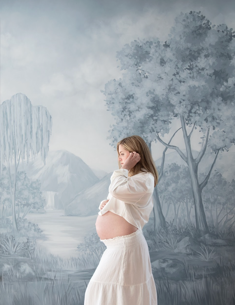 The founder of The Postpartum Village Rachel Bernhard holds her pregnant stomach against a blue forest wallpaper.