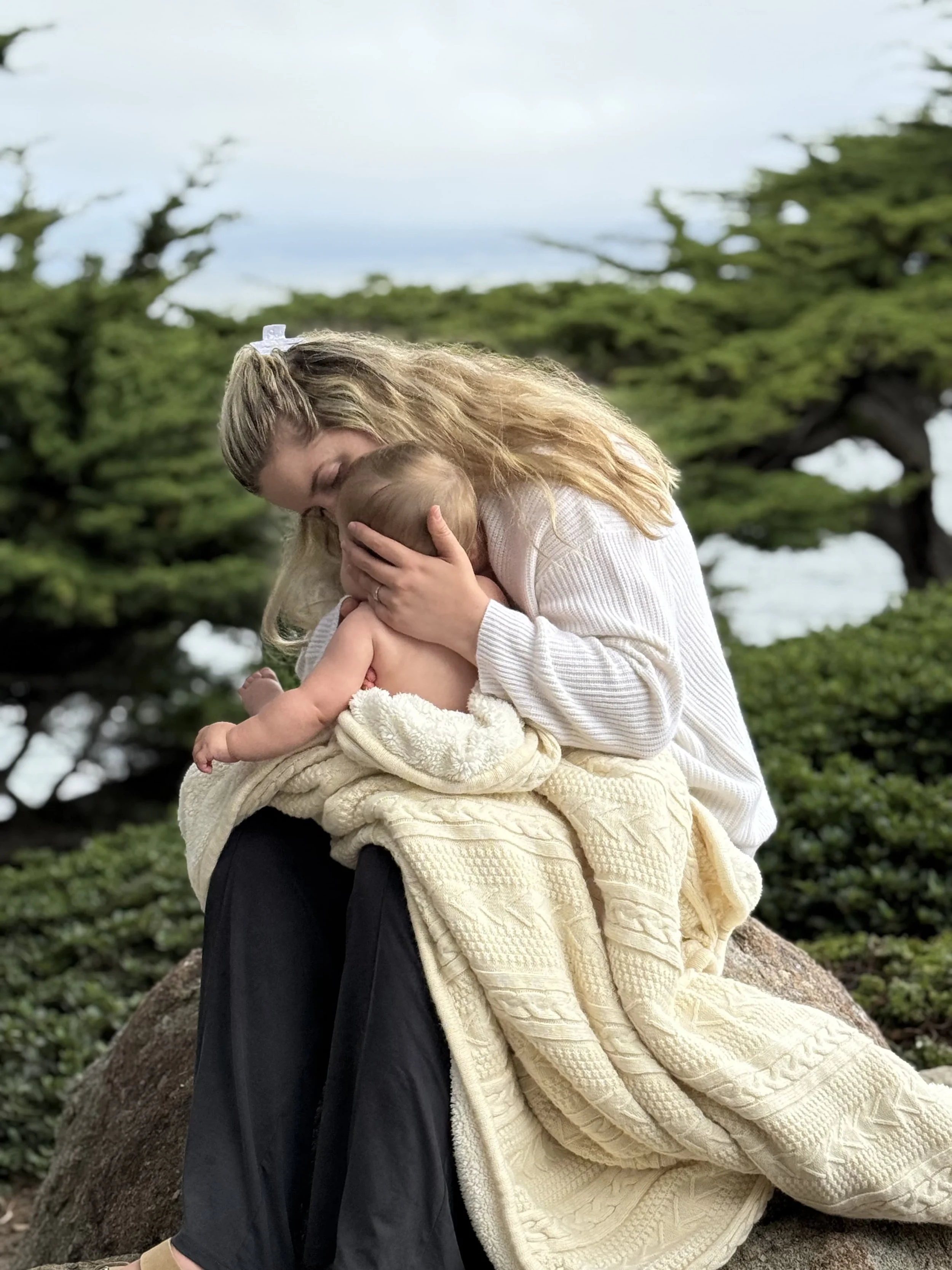 Rachel Bernhard, virtual and holistic postpartum support group founder, holds her newborn in a yellow blanket outside.