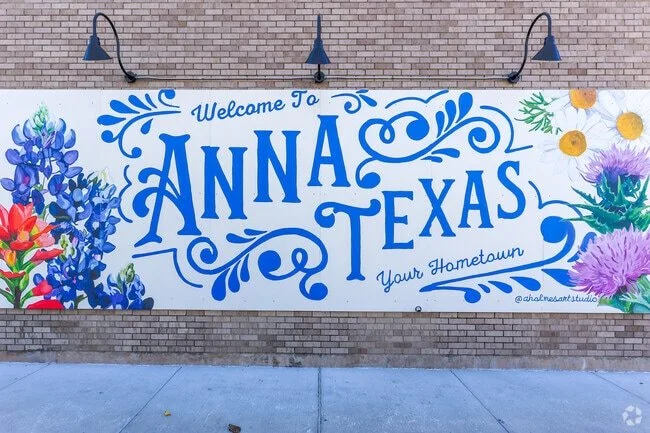 Colorful mural on a brick wall that says "Welcome to Anna Texas, Your Hometown," decorated with blue and purple flowers.