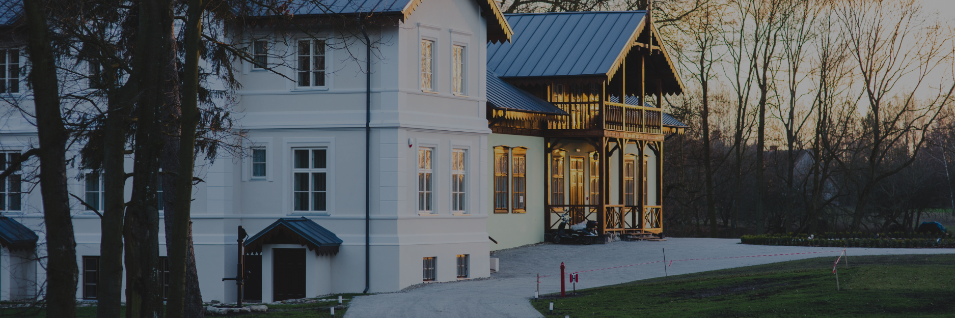 A large white house with a wooden balcony and dark blue metal roof, surrounded by leafless trees at sunset.