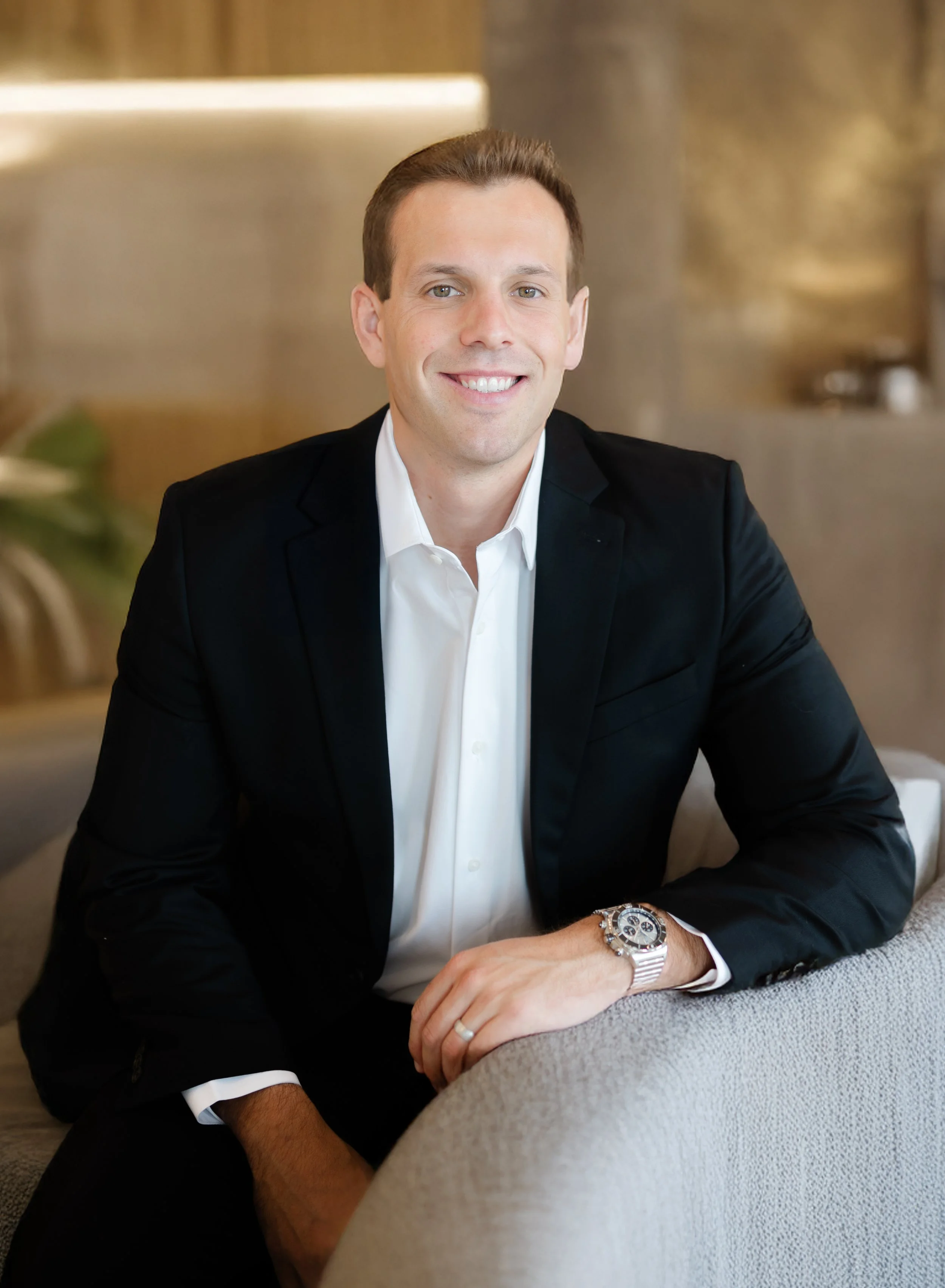A smiling man with brown hair, wearing a black blazer, white shirt, and a large watch, sitting indoors with a blurred background.