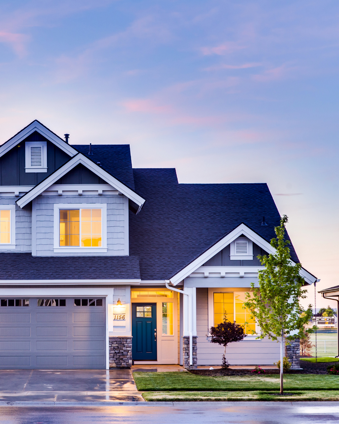 A two-story suburban house with a dark blue roof, white and gray siding, a front porch with a teal door, lit windows, a manicured lawn, small trees, and a wet driveway at dusk.