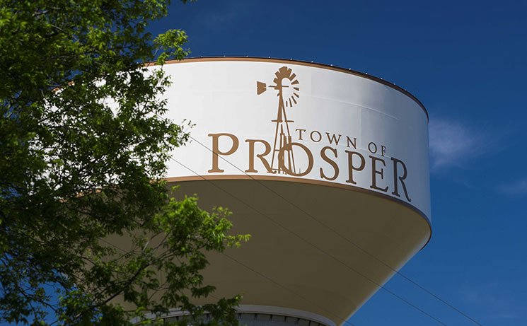 Water tower with the town of Prosper logo, featuring a windmill, against a blue sky with some green tree branches in the foreground.