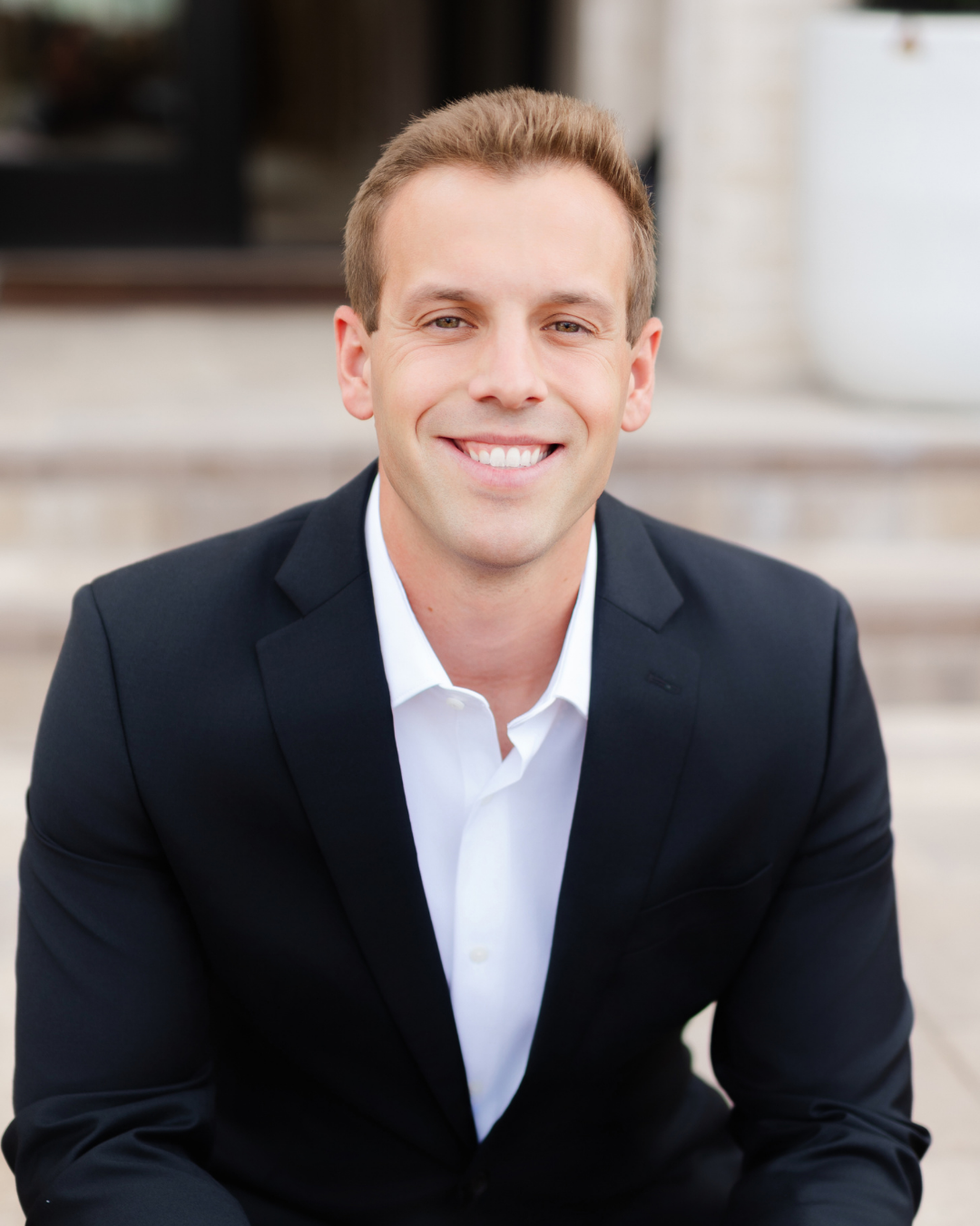A young man in a black suit and white shirt smiling outdoors in front of a light-colored building with steps.