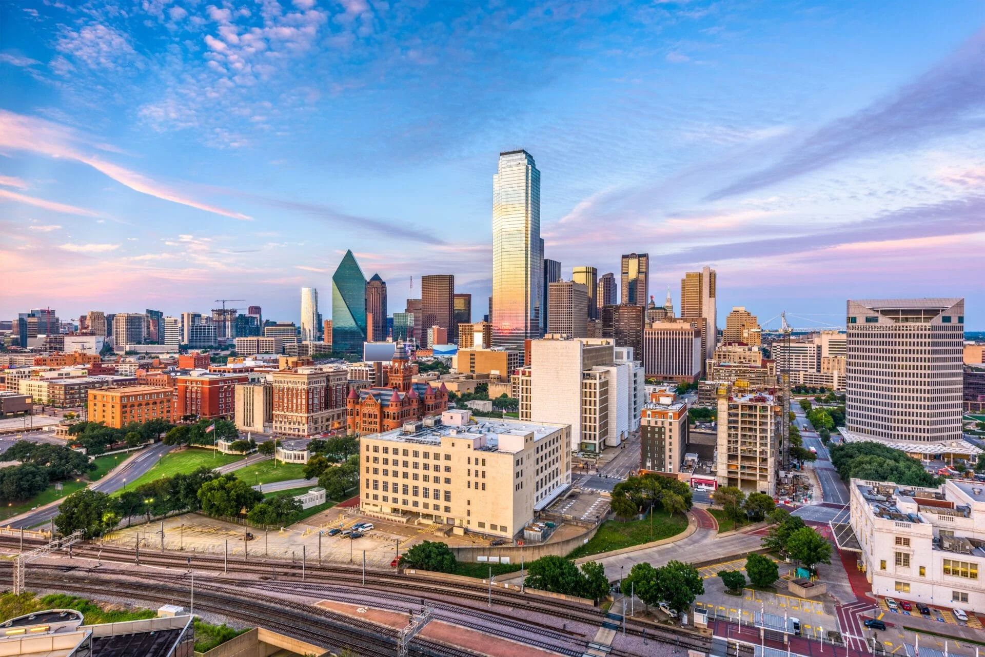 A city skyline with tall skyscrapers and a mix of modern and historic buildings under a colorful, partly cloudy sky during sunset.