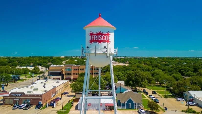 Water tower with 'Frisco' sign above a small town with buildings, parked cars, and lush green trees under a clear blue sky.