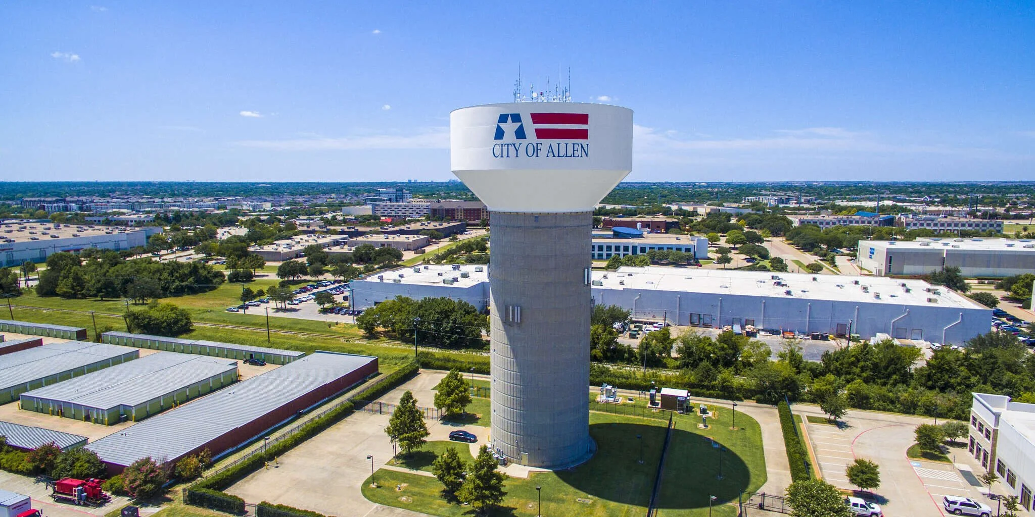 A tall water tower with the logo and words 'City of Allen' on it, located in an urban area with buildings, trees, and parking lots, under a clear blue sky.