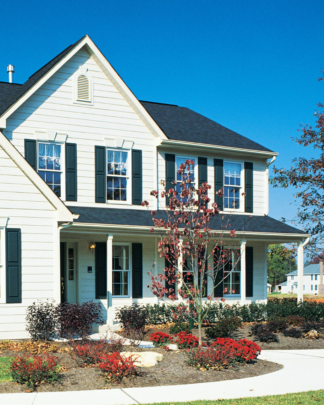 A two-story white house with black shutters and a dark roof, surrounded by a yard with red and dark purple bushes, a young red-leafed tree, and a sidewalk, under a clear blue sky.
