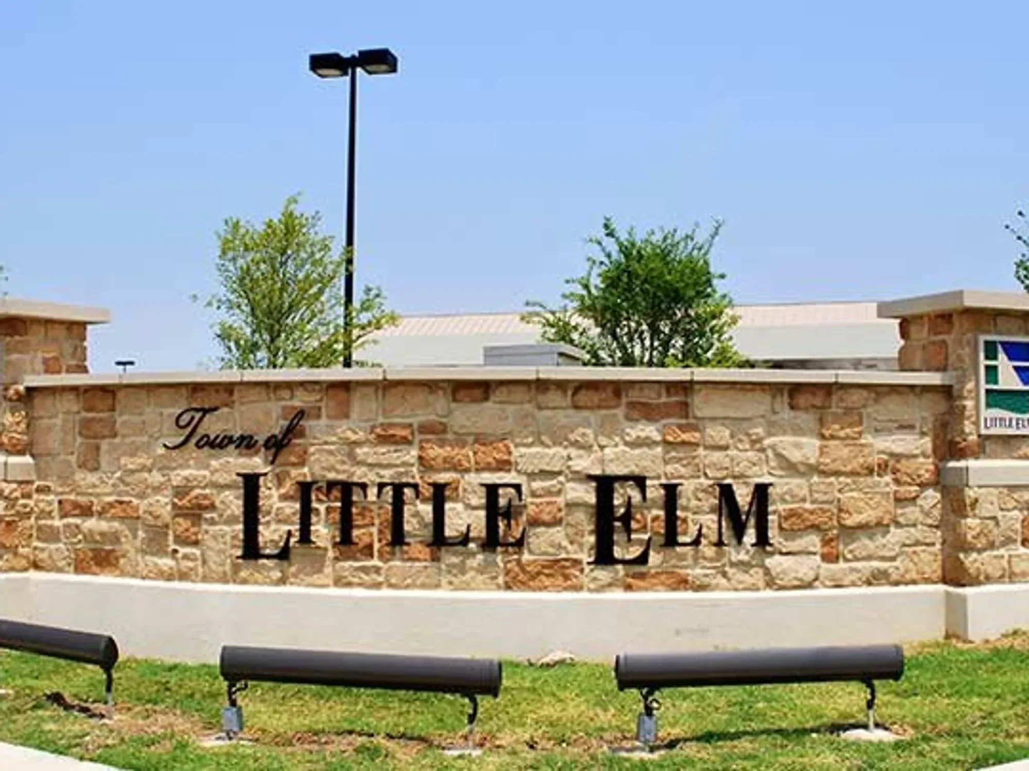 A brick sign with black lettering reads 'Town of Little Elm' against a background of trees, a building roof, and a clear blue sky. There are two black benches in the foreground.