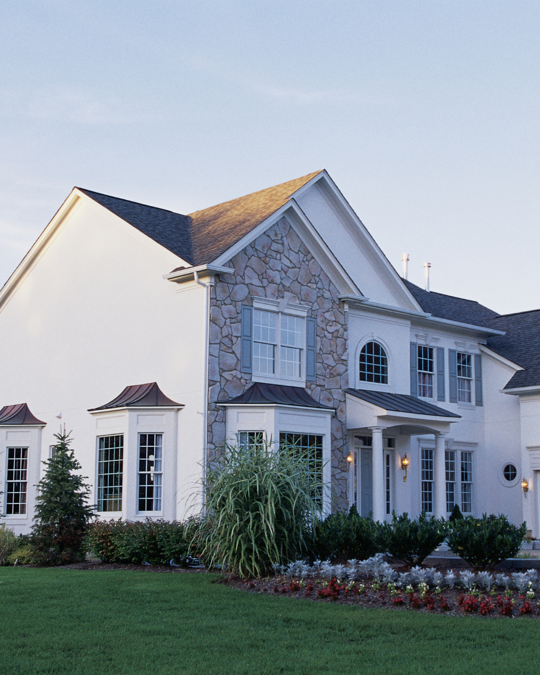 A large, elegant white house with stone accents, multiple windows, and a well-maintained lawn with shrubs and flowers. External lights are on near the entrance.