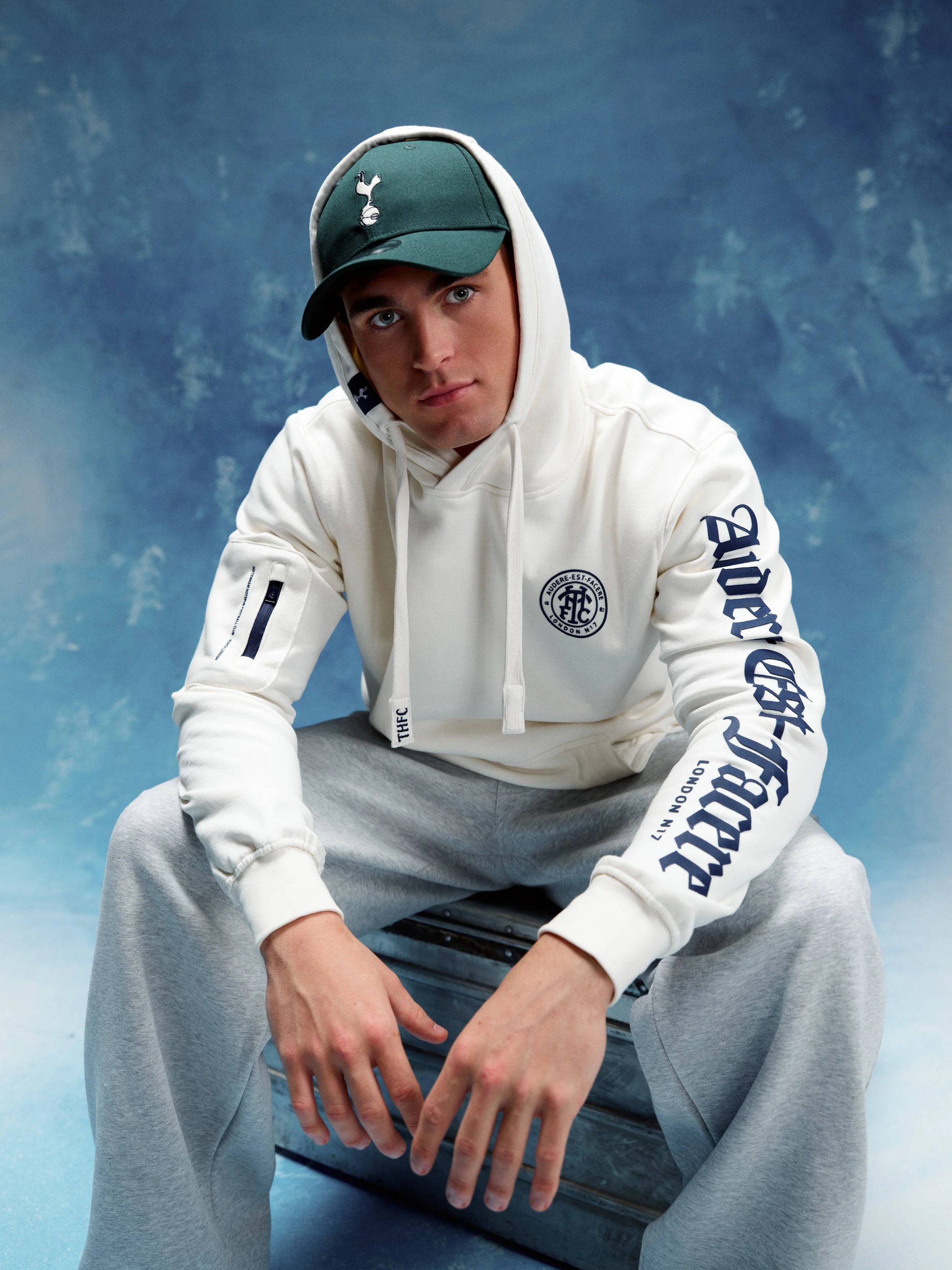 A young man wearing a white hoodie with blue text on the sleeves, grey sweatpants, and a green baseball cap tilted to the side, sitting on a silver metal box against a blue background.