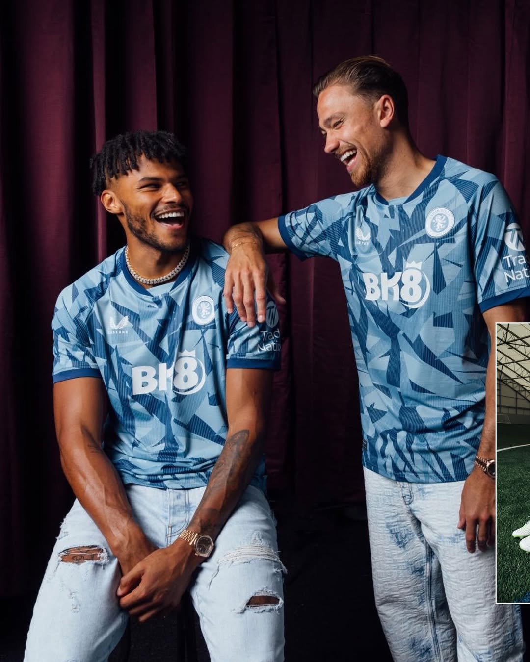 Two men in blue sports jerseys laughing and smiling, one has tattoos and a necklace, the other has a watch and light-colored ripped jeans, standing against a purple curtain background.