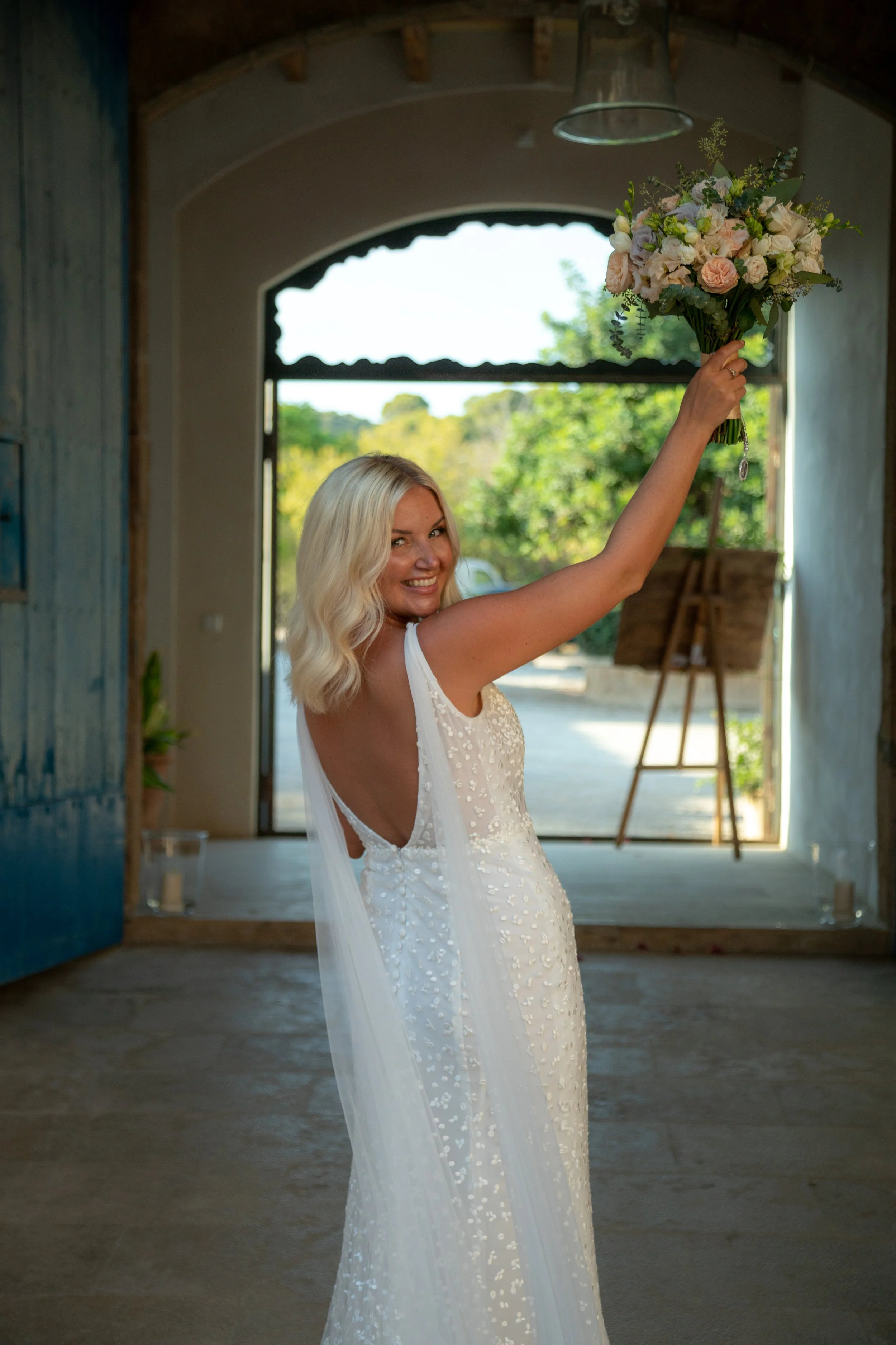 A woman in a white wedding dress holding a bouquet of flowers, smiling inside a rustic building with an open door and greenery outside.