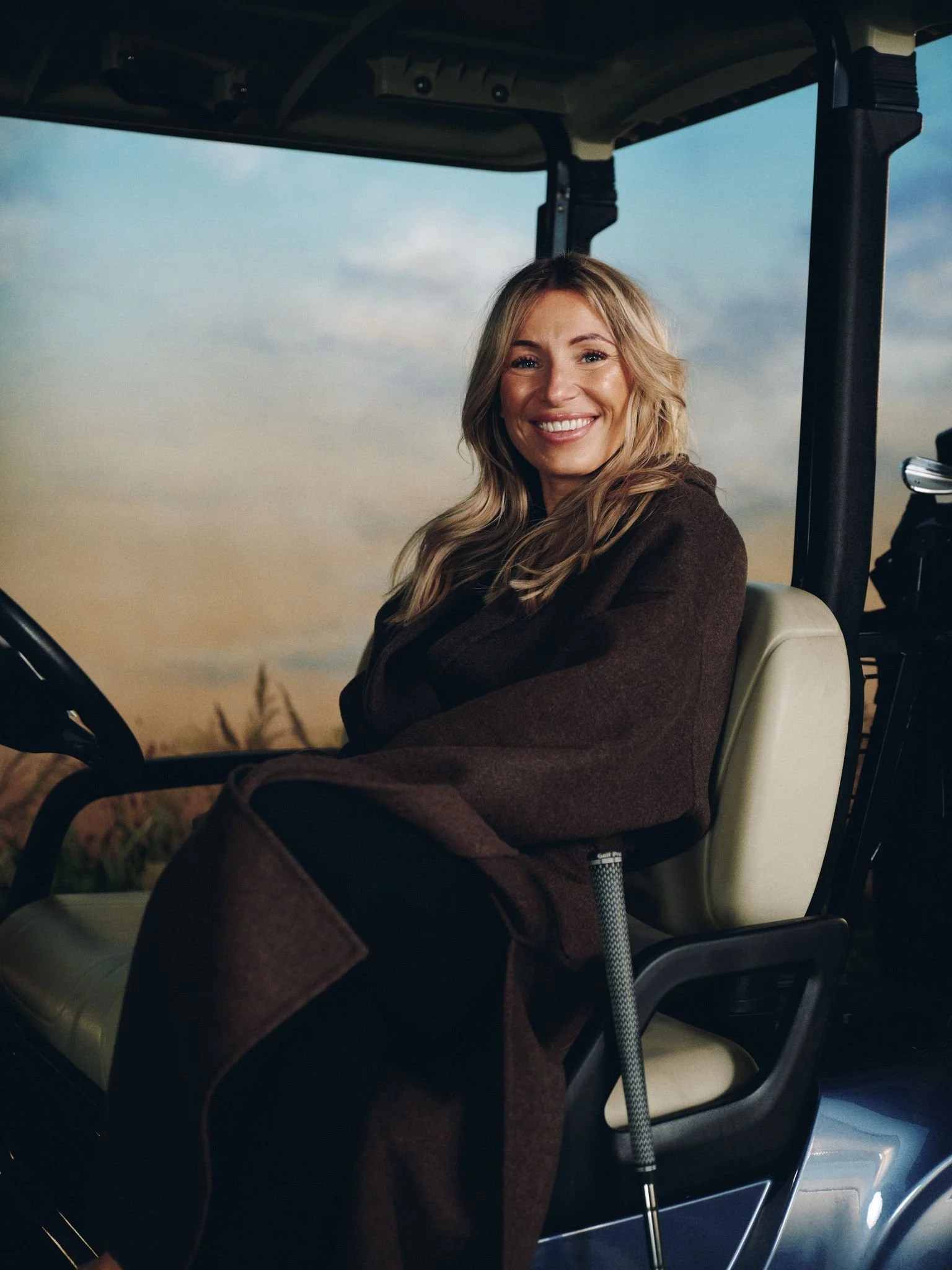 A woman sitting in a golf cart, smiling at the camera, with a background of a cloudy sky and field.