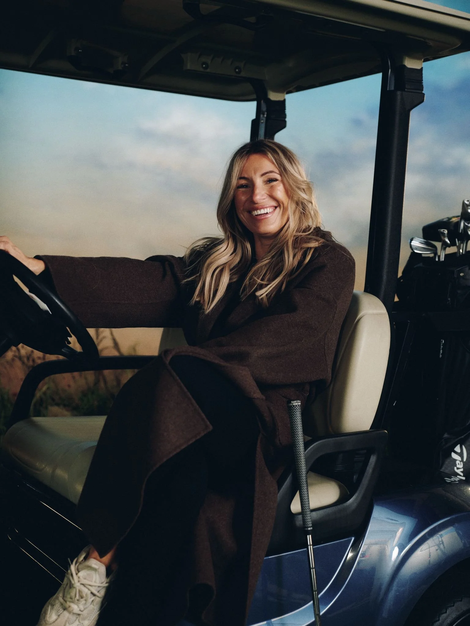 A smiling woman sitting in a golf cart, with golf clubs behind her, outdoors during late afternoon or early evening.