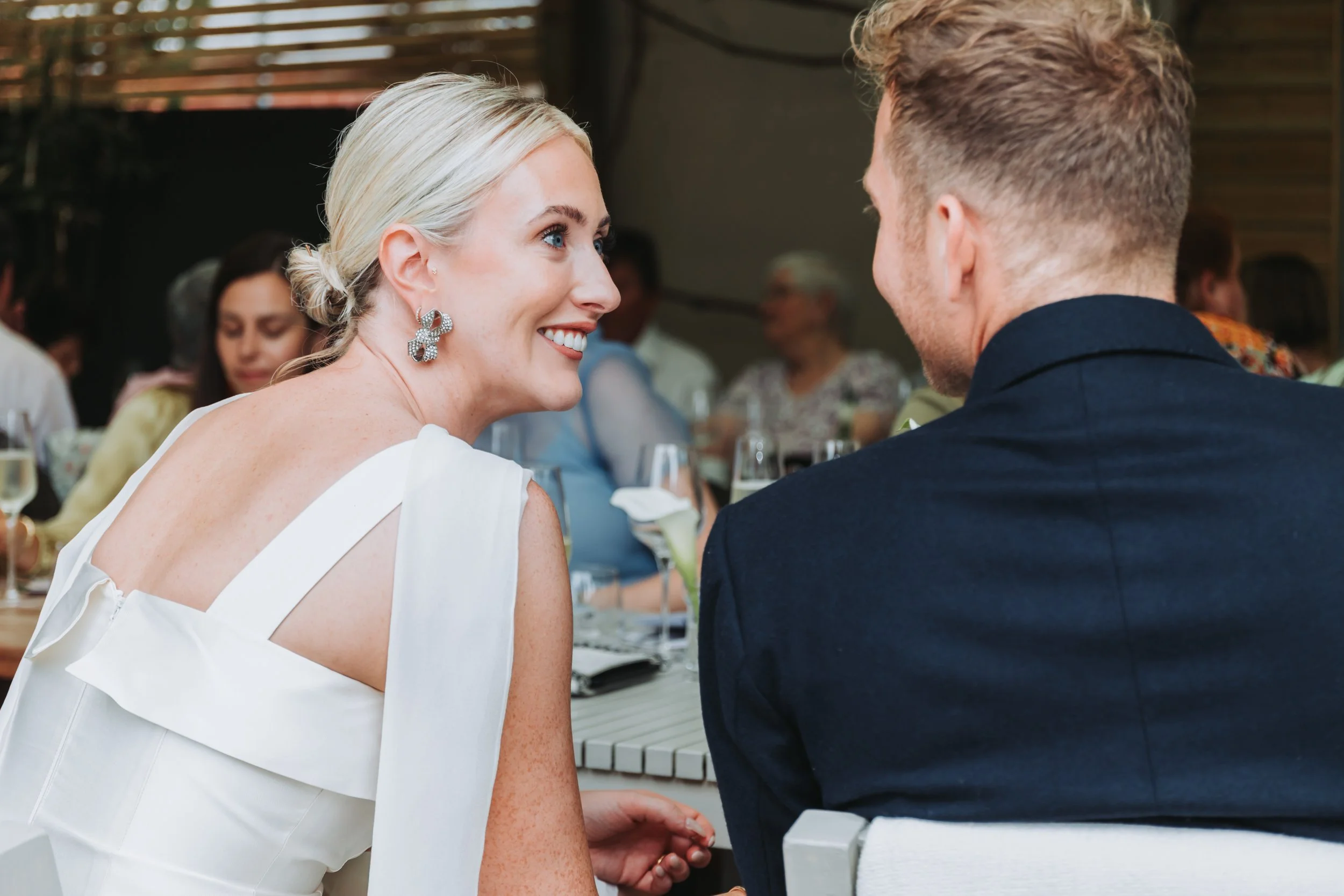 A woman with blonde hair and earrings smiling at a man during a social gathering or celebration, with other people and drinks in the background.