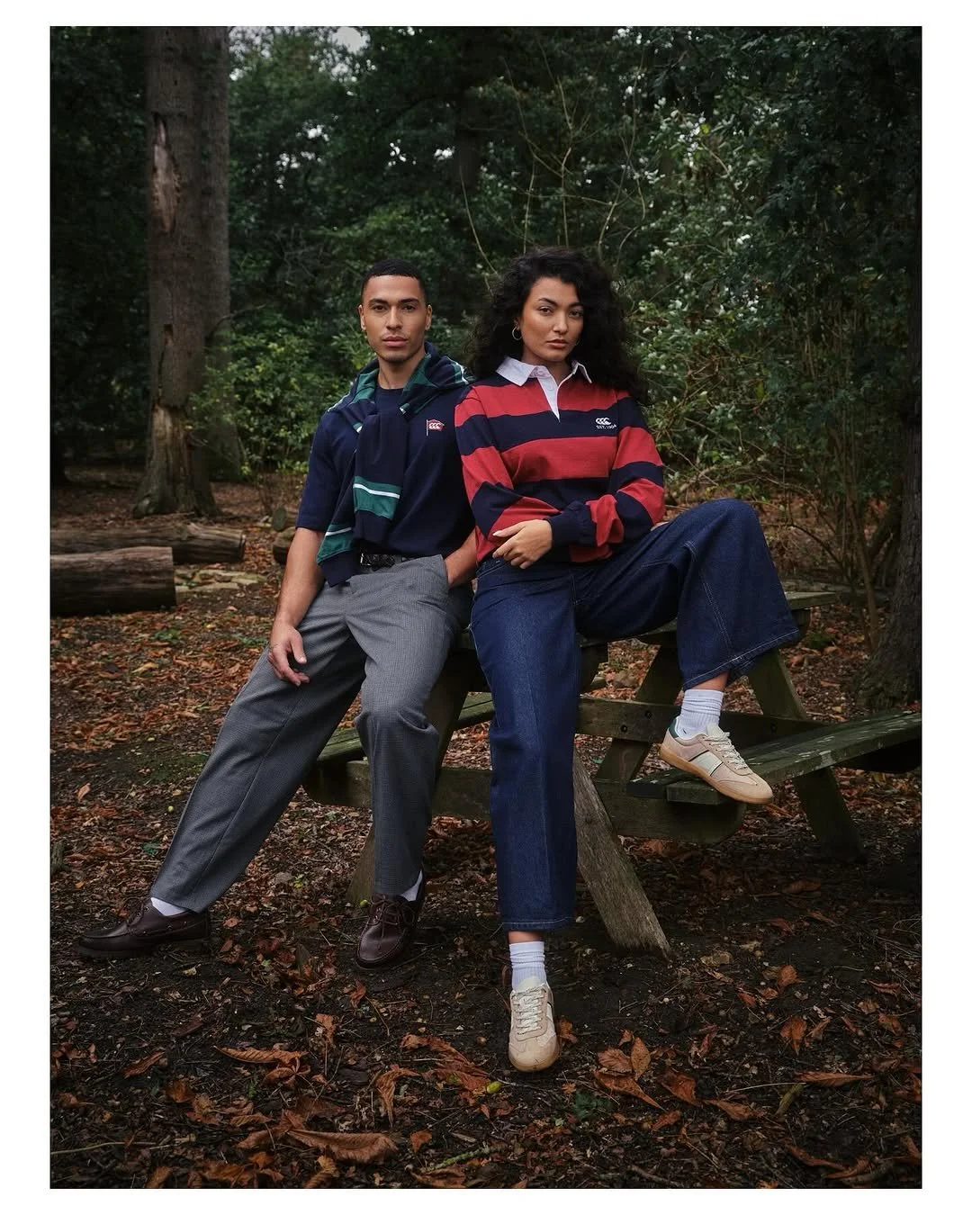 Two young adults sitting on a wooden bench in a forest, dressed casually in rugby shirts and jeans.