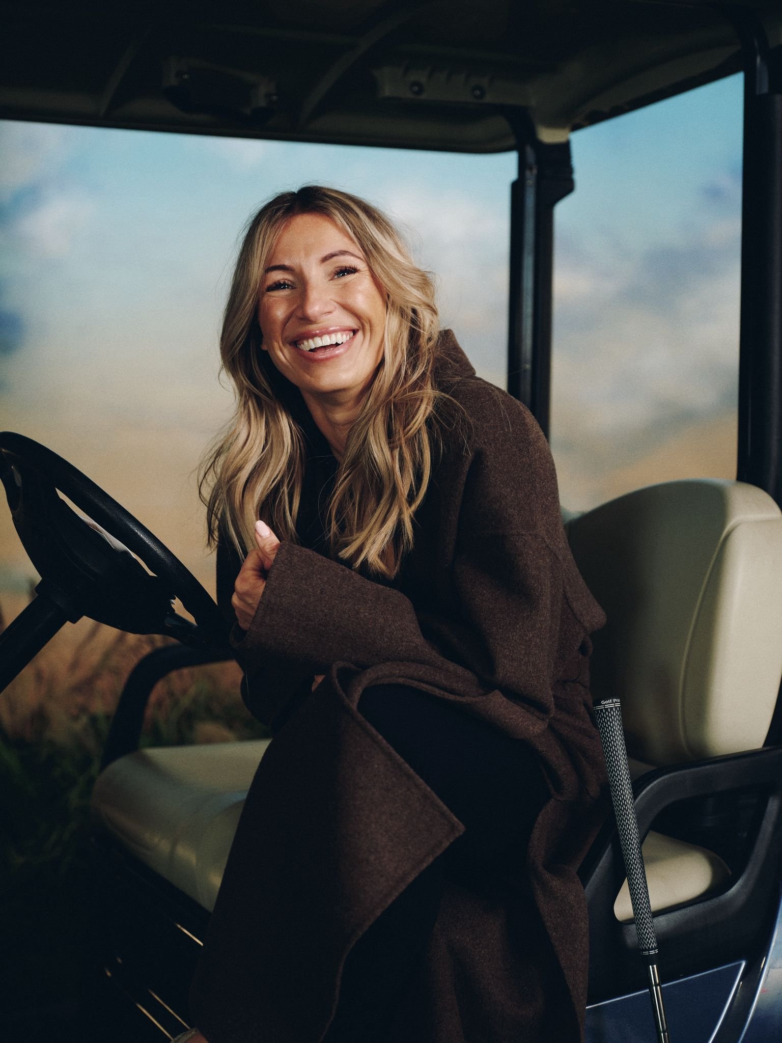 A woman with long blonde hair smiling while sitting in a golf cart, holding the steering wheel, with a stick golf club leaning against the seat.