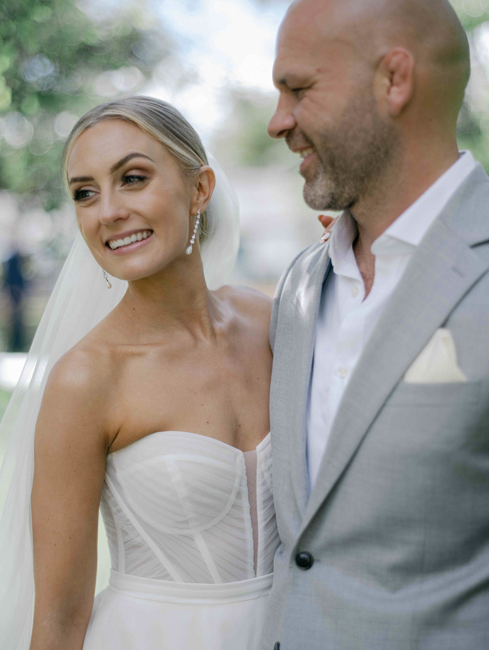 A bride and groom are smiling and standing close to each other outdoors during their wedding. The bride is wearing a strapless white wedding dress with a sheer bodice and long earrings. The groom is in a light gray suit with a white shirt.