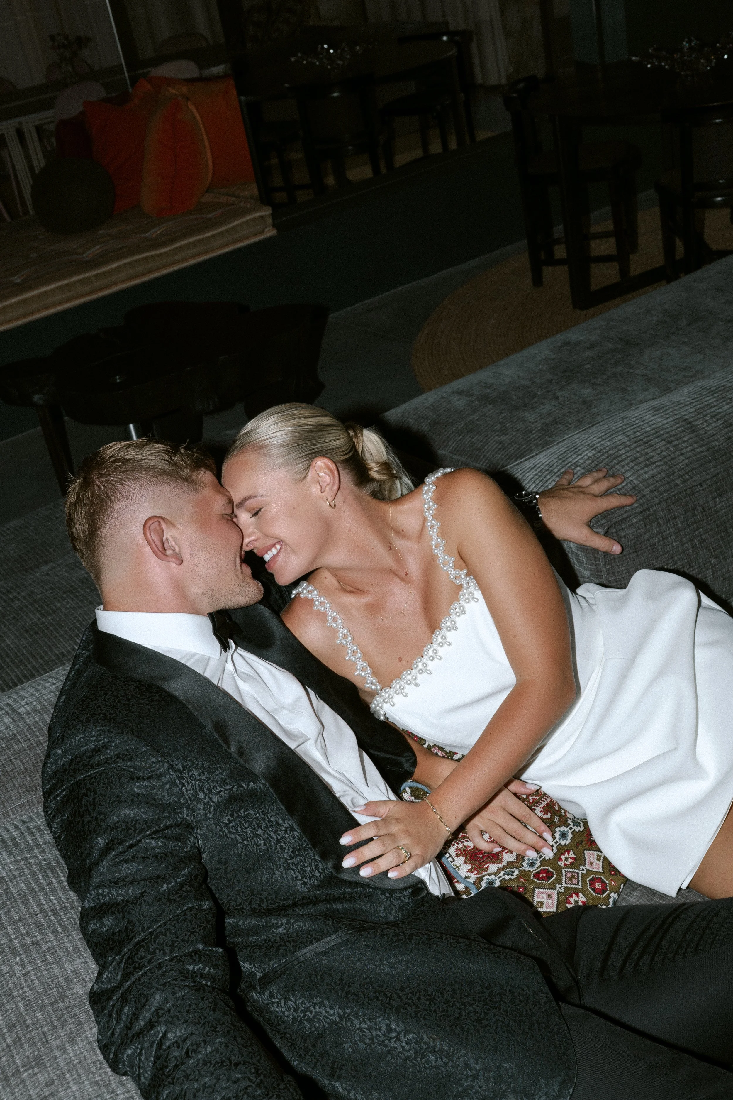A smiling couple dressed in formal attire, lying close together on a sofa, about to kiss in a cozy, dimly lit indoor setting.