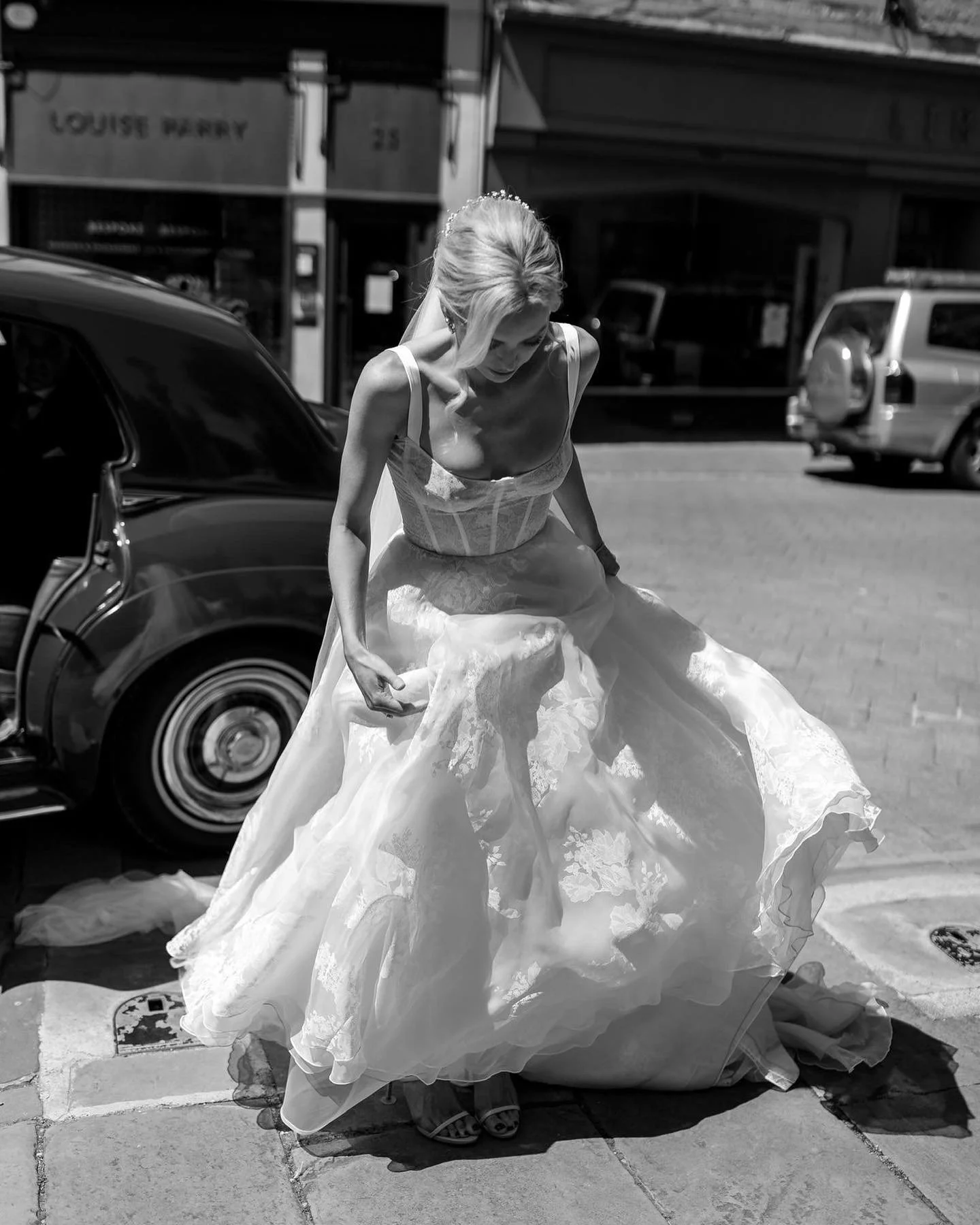 A woman in a wedding dress stepping down from a curb on a city street, with cars and storefronts in the background.
