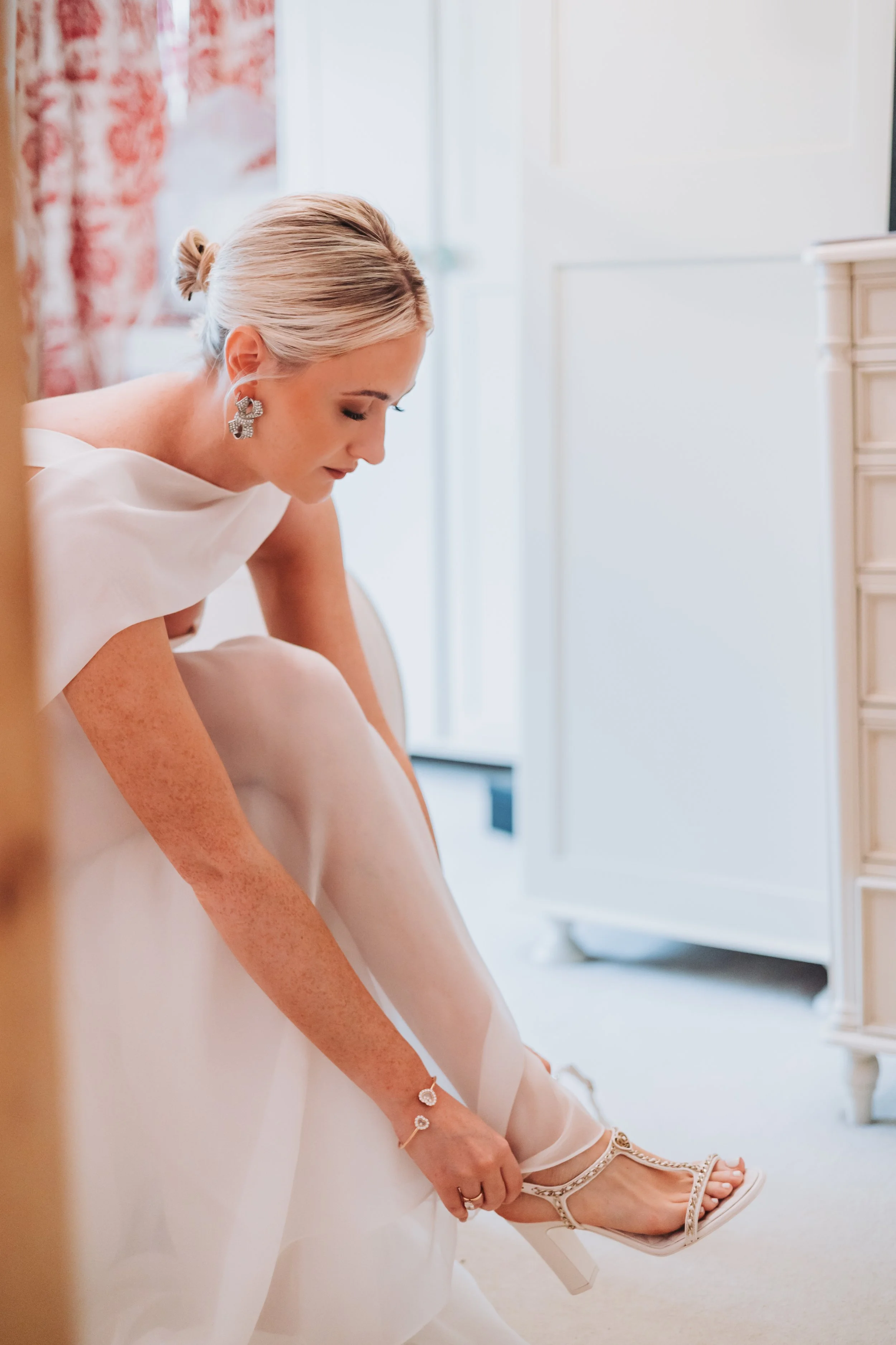 A woman in a white wedding dress is bending down to put on her white high-heeled sandal shoe.