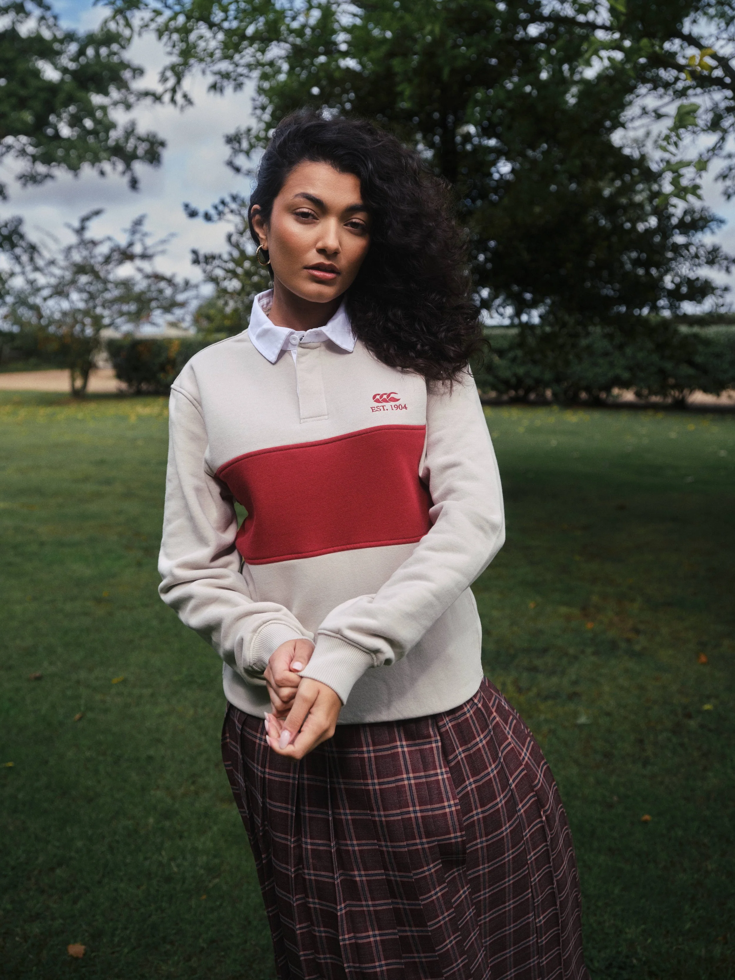 A woman with curly black hair outdoors, wearing a beige and red sweatshirt over a white collared shirt and a plaid skirt.