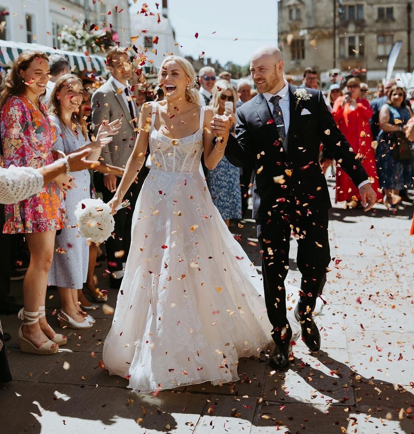 A newlywed couple walking amid confetti, holding hands and smiling, surrounded by friends and family on a sunny day.