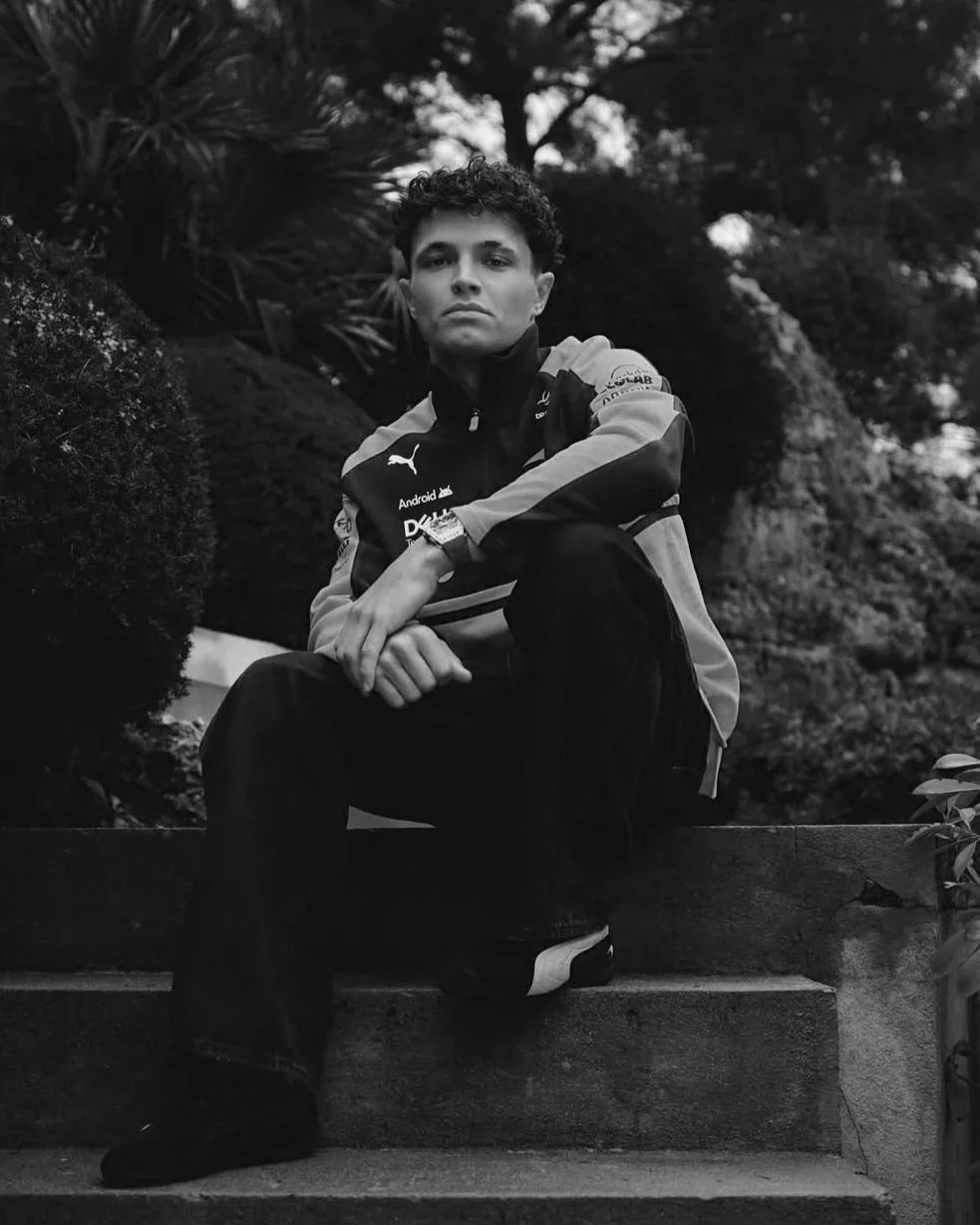 A young man with curly hair sitting on outdoor stairs, wearing a sports jacket and dark pants, surrounded by trees and foliage in a park setting.