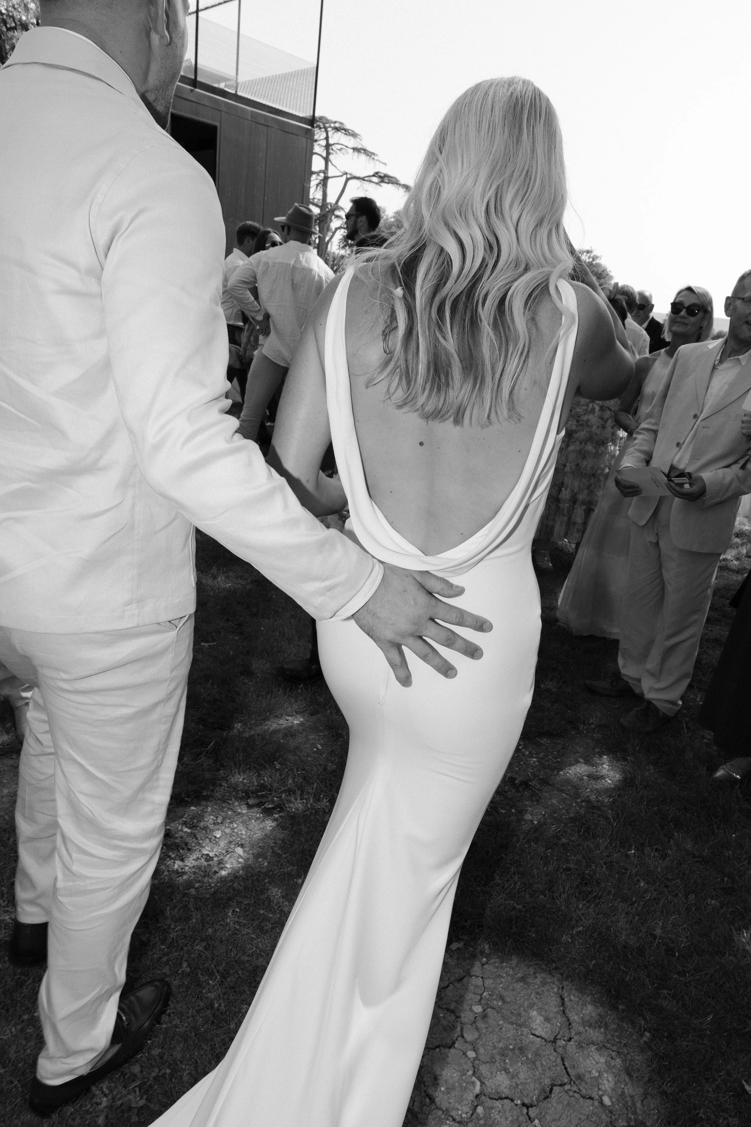 A woman in a backless white dress, standing outdoors at a social gathering, with a man in a white suit touching her waist. Multiple people are in the background, some wearing sunglasses and light-colored clothing.