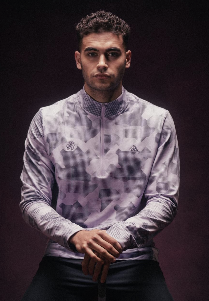 A young man with short, curly dark hair and a serious expression, wearing a light purple and gray camouflage-patterned Nike sports jacket with a zipper, standing against a dark background.