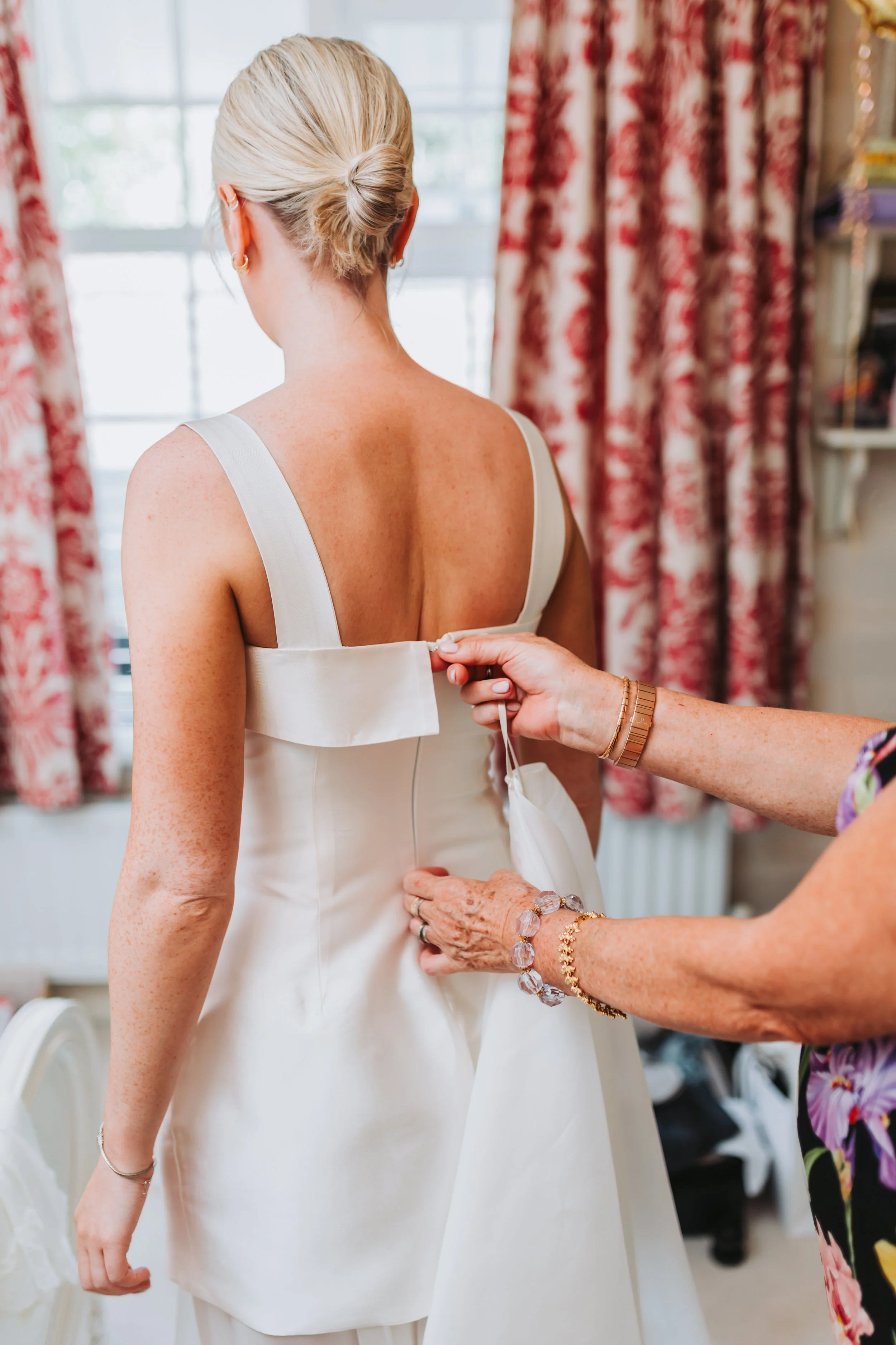 A bride in a white wedding dress is being assisted with her gown by another woman, who is fastening the back of the dress. The scene appears to be in a well-lit room with floral curtains and a bookshelf in the background.