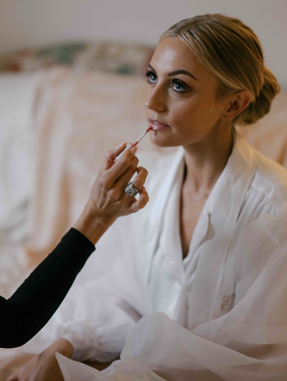 A woman with blonde hair and blue eyes getting her makeup or lipstick done by another person with painted nails, indoors with a blurred background.