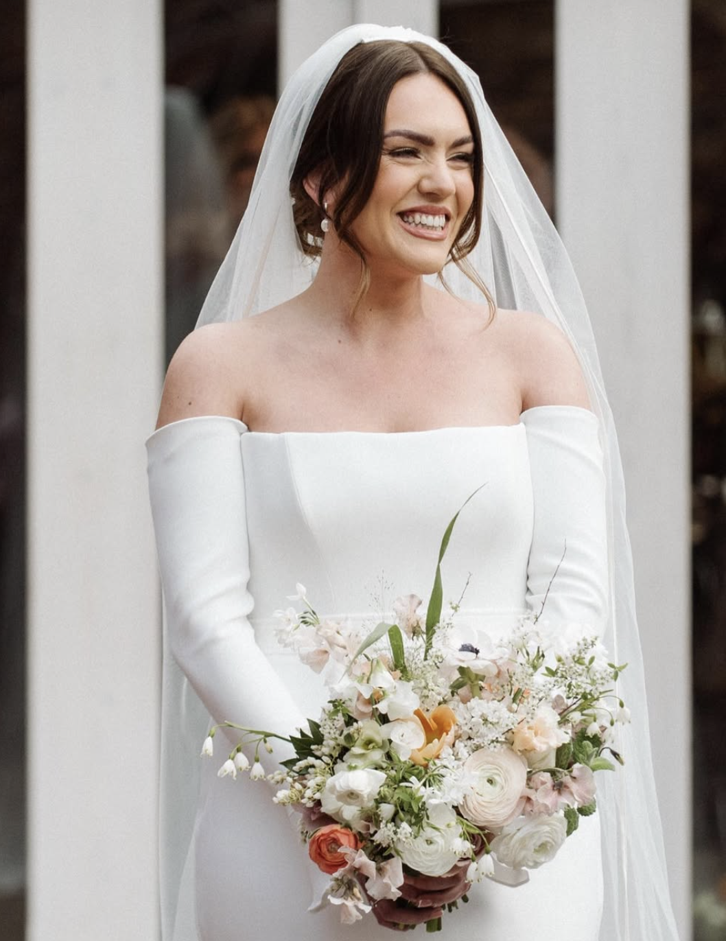 A bride in an off-shoulder white wedding dress holding a bouquet of flowers, smiling with eyes closed, wearing pearl earrings and a veil.