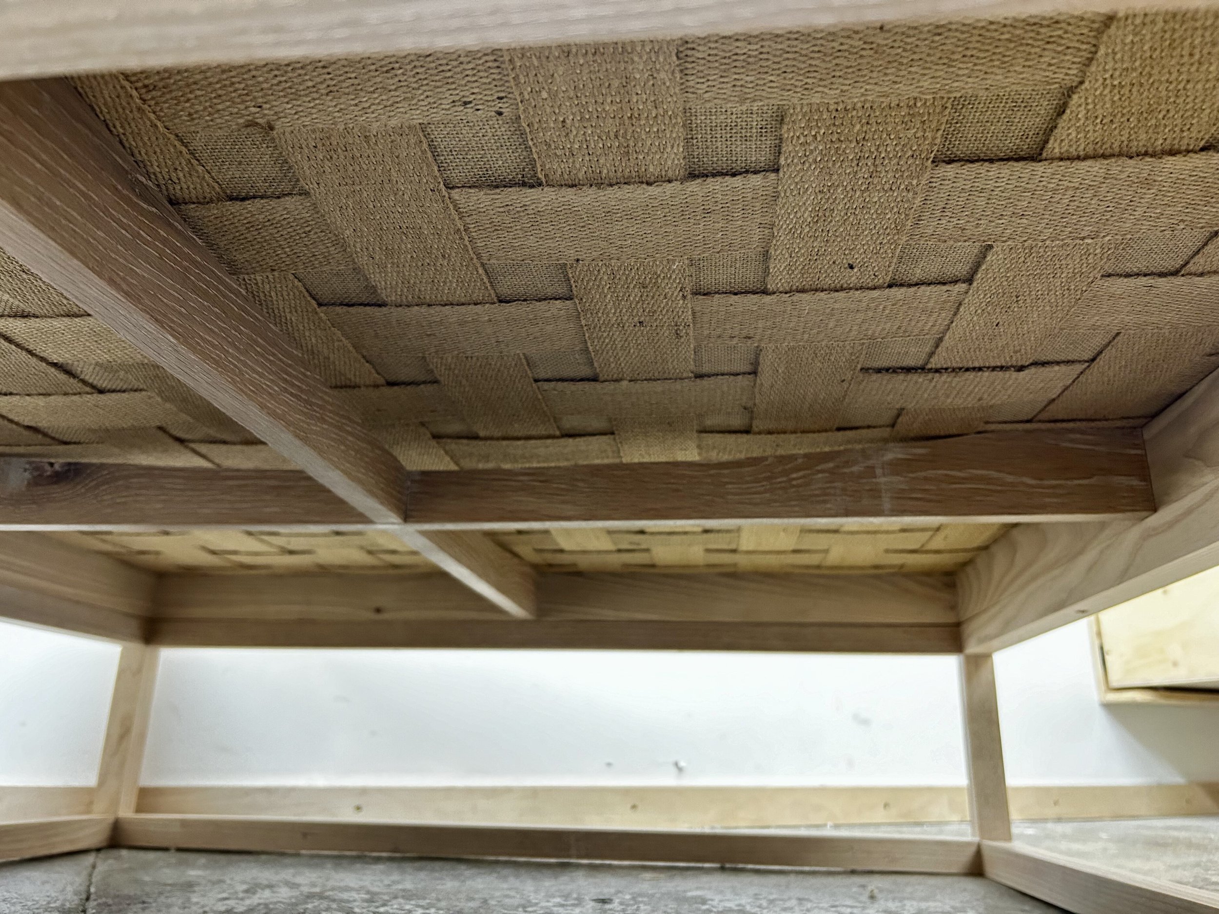 The underside of a wooden table with a woven wicker bottom shelf and wooden support beams.