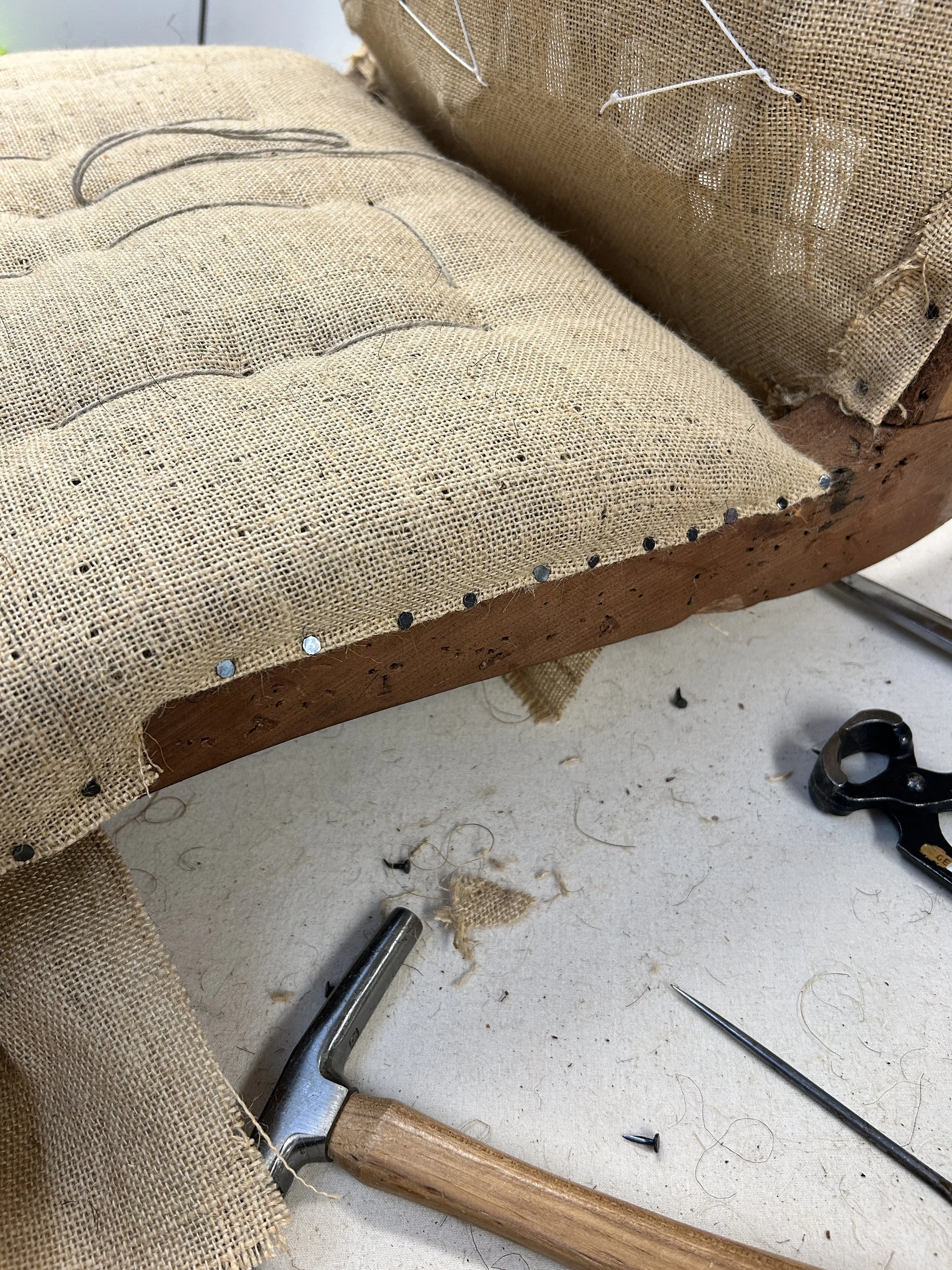A piece of hessian tacked onto a wooden frame with upholstery tools, including a hammer, regulator and needle, on a white surface with fabric scraps.