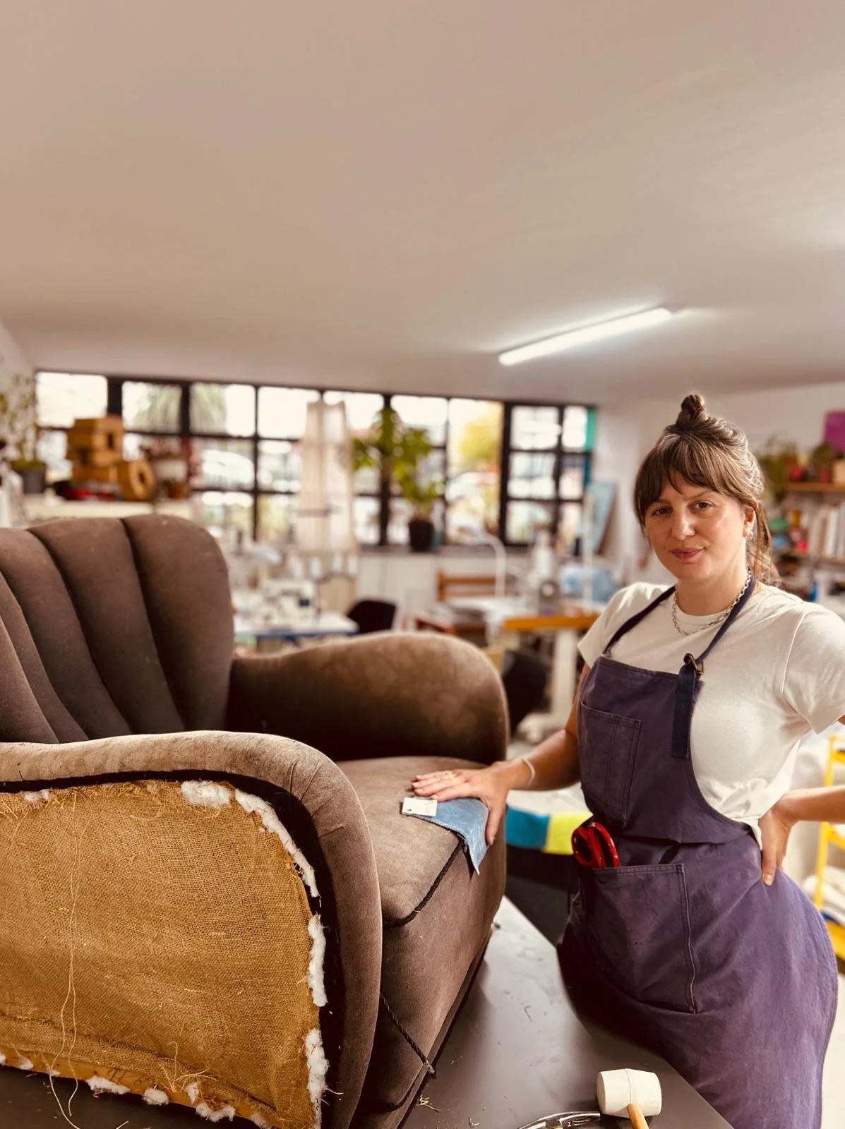 A young woman wearing a white t-shirt and dark apron is working on reupholstering a vintage brown armchair in a workshop, with shelves, plants, and windows in the background.