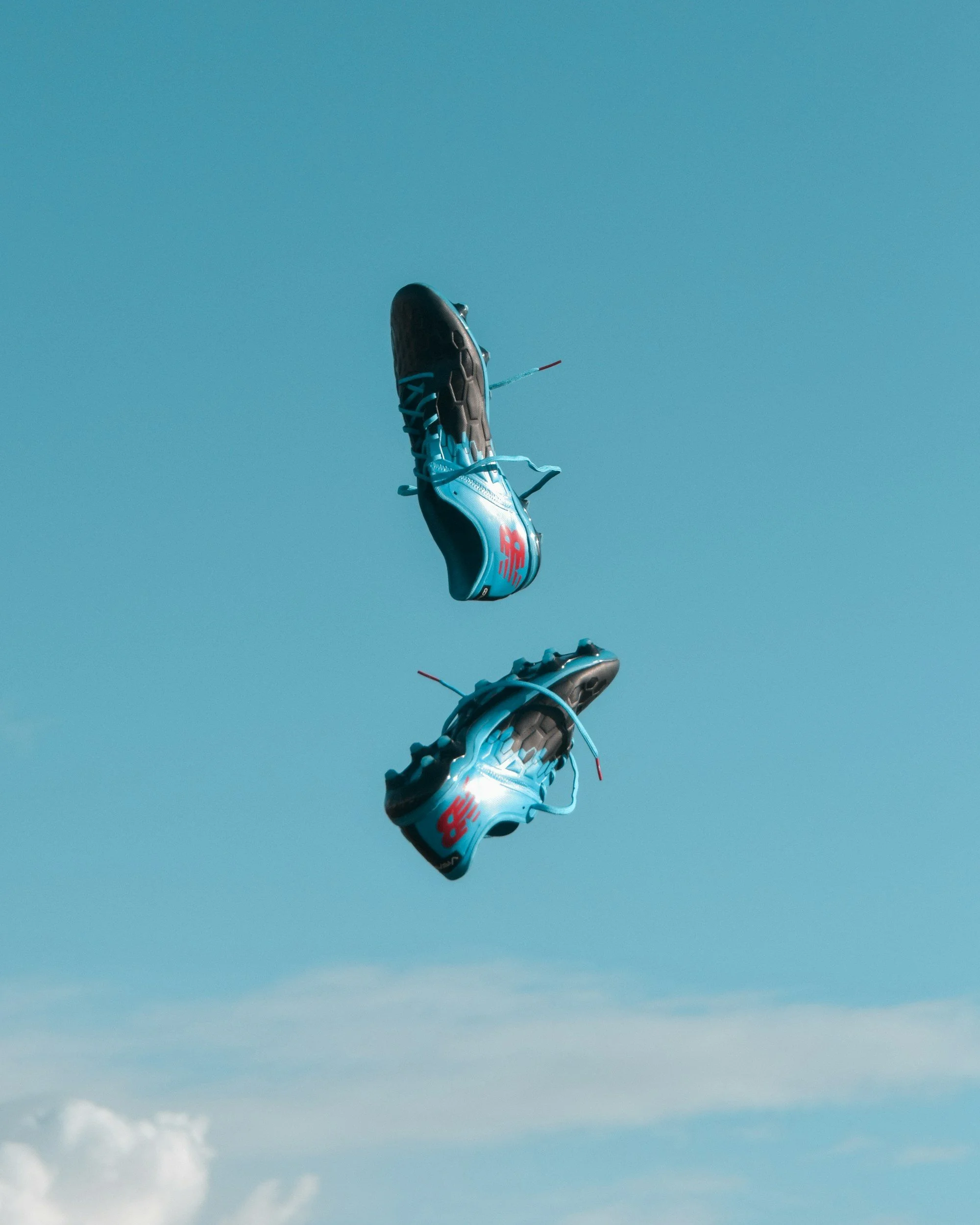 Two racing shoes suspended in the air against a blue sky with a small cloud below.