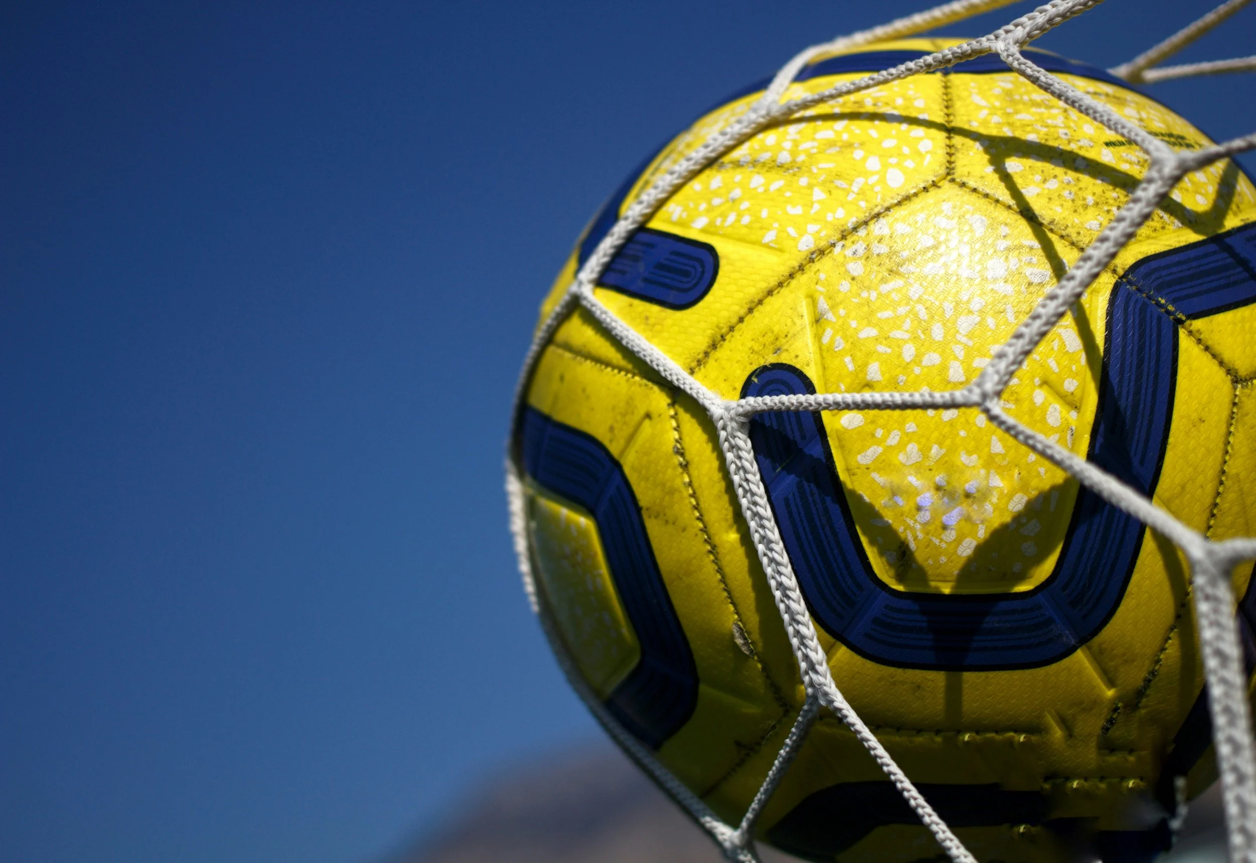 Yellow and blue soccer ball in a white net against a blue sky background.
